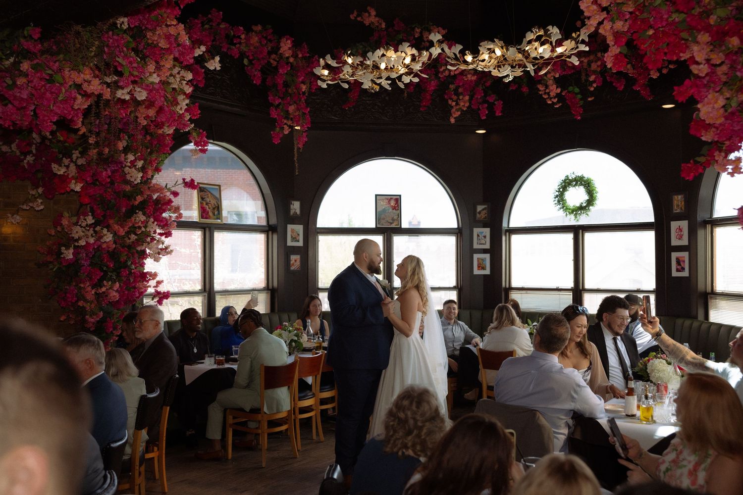 Bride and groom stand together in elegant restaurant reception hall adorned with vibrant pink floral ceiling decorations.