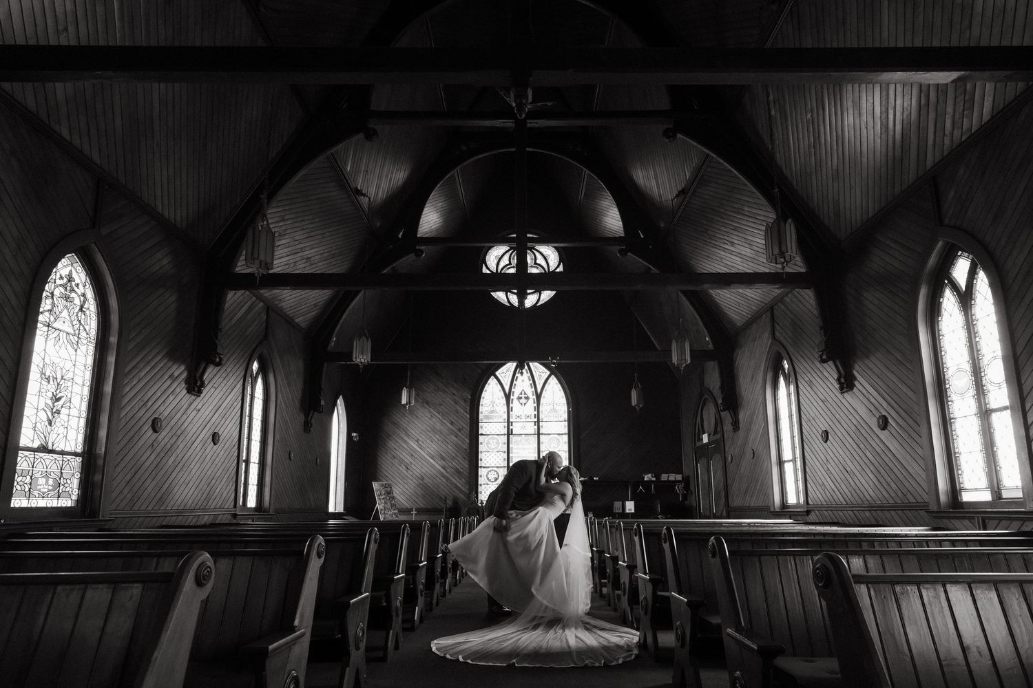 Black and white photo of bride and groom sharing a kiss in a stunning wooden church with arched windows.
