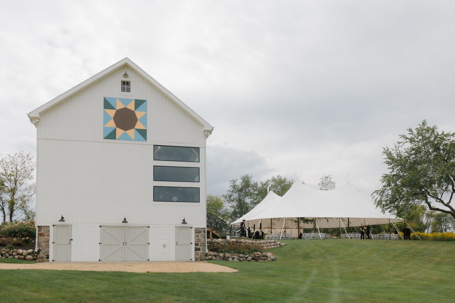 White barn with colorful quilt square and large white wedding tent on a green lawn.