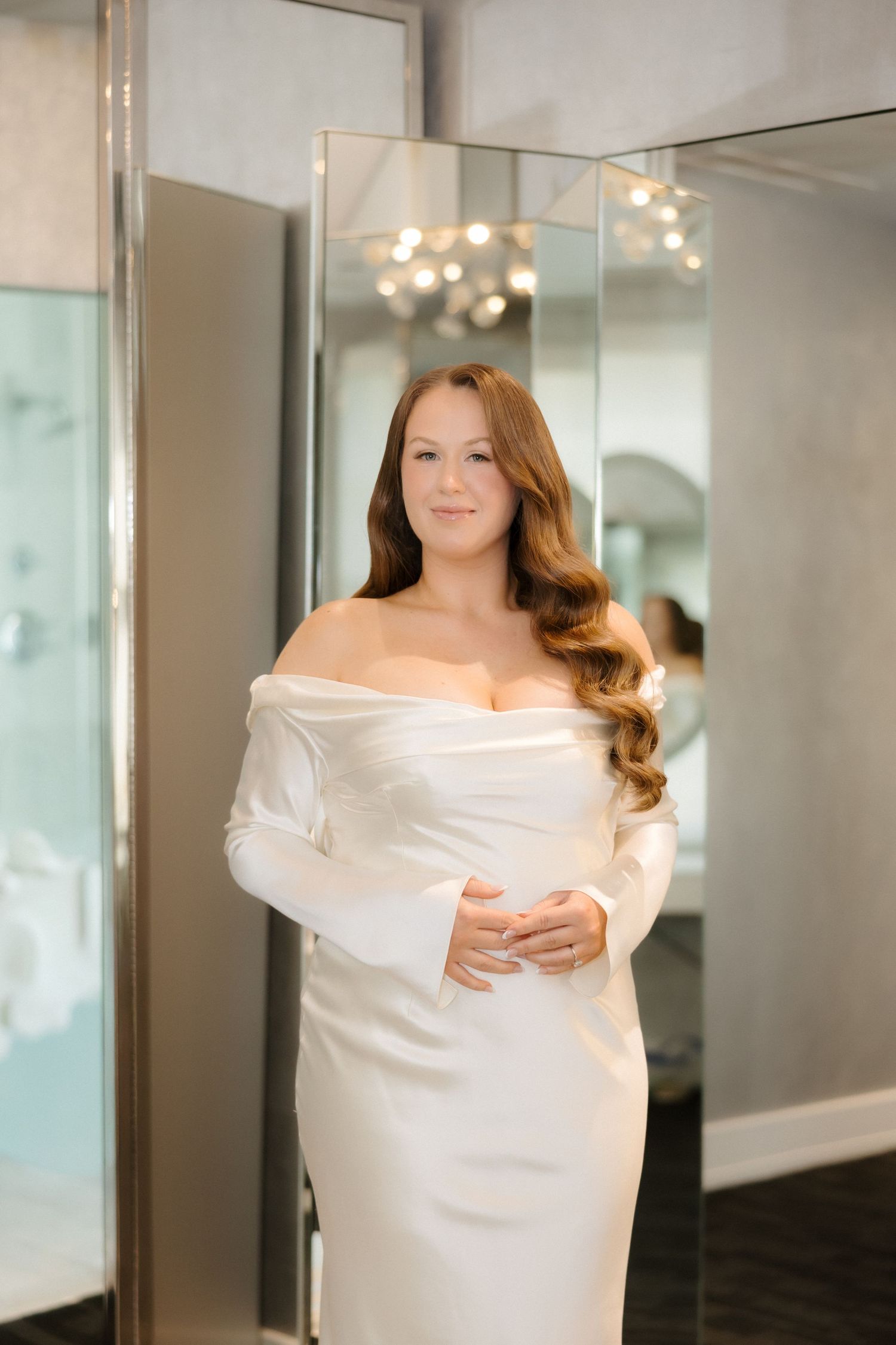 Bride wearing an elegant off-shoulder white satin gown with long sleeves, posing in a bridal boutique fitting room.