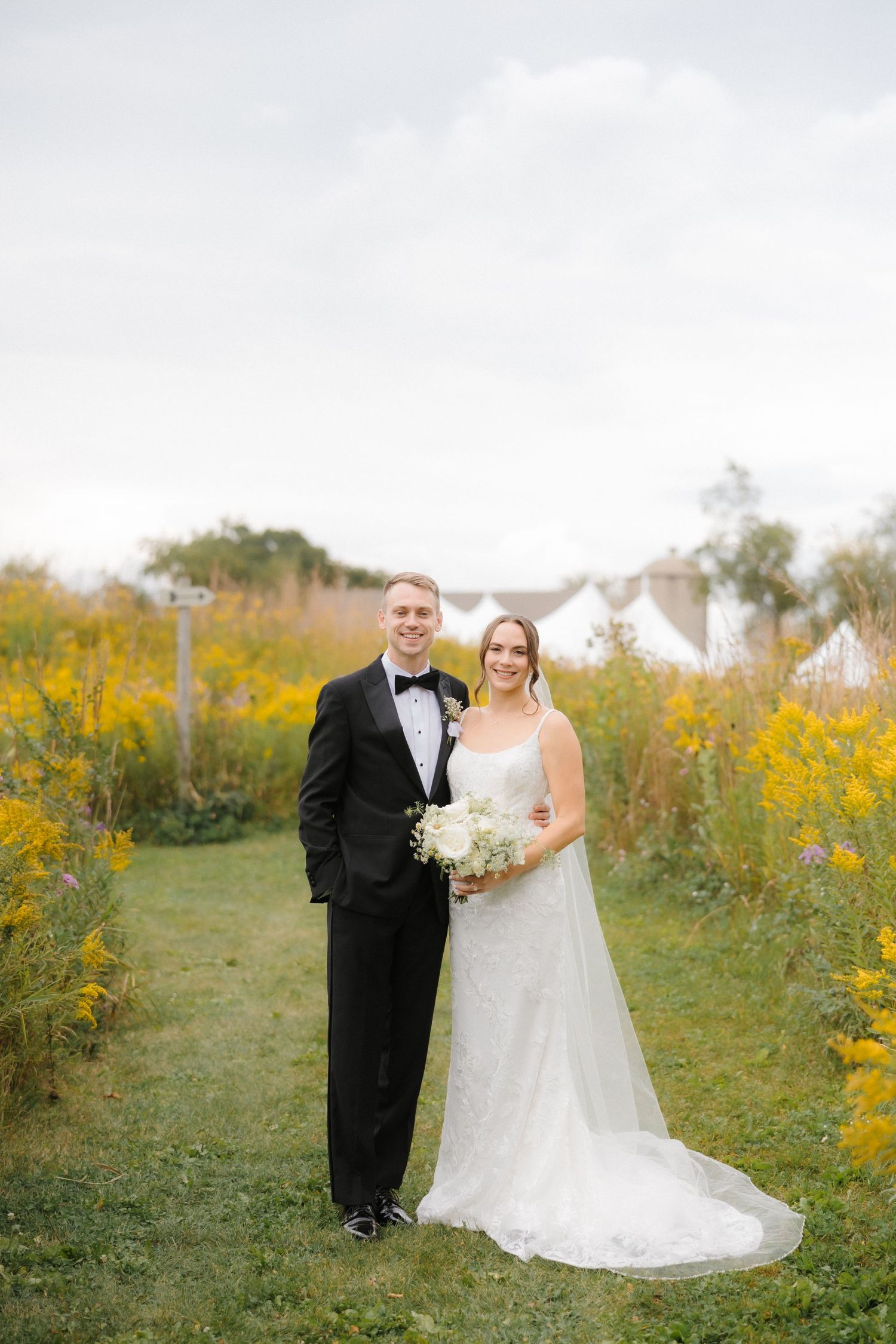 Bride and groom posing together in yellow wildflower field with white wedding tent in background.