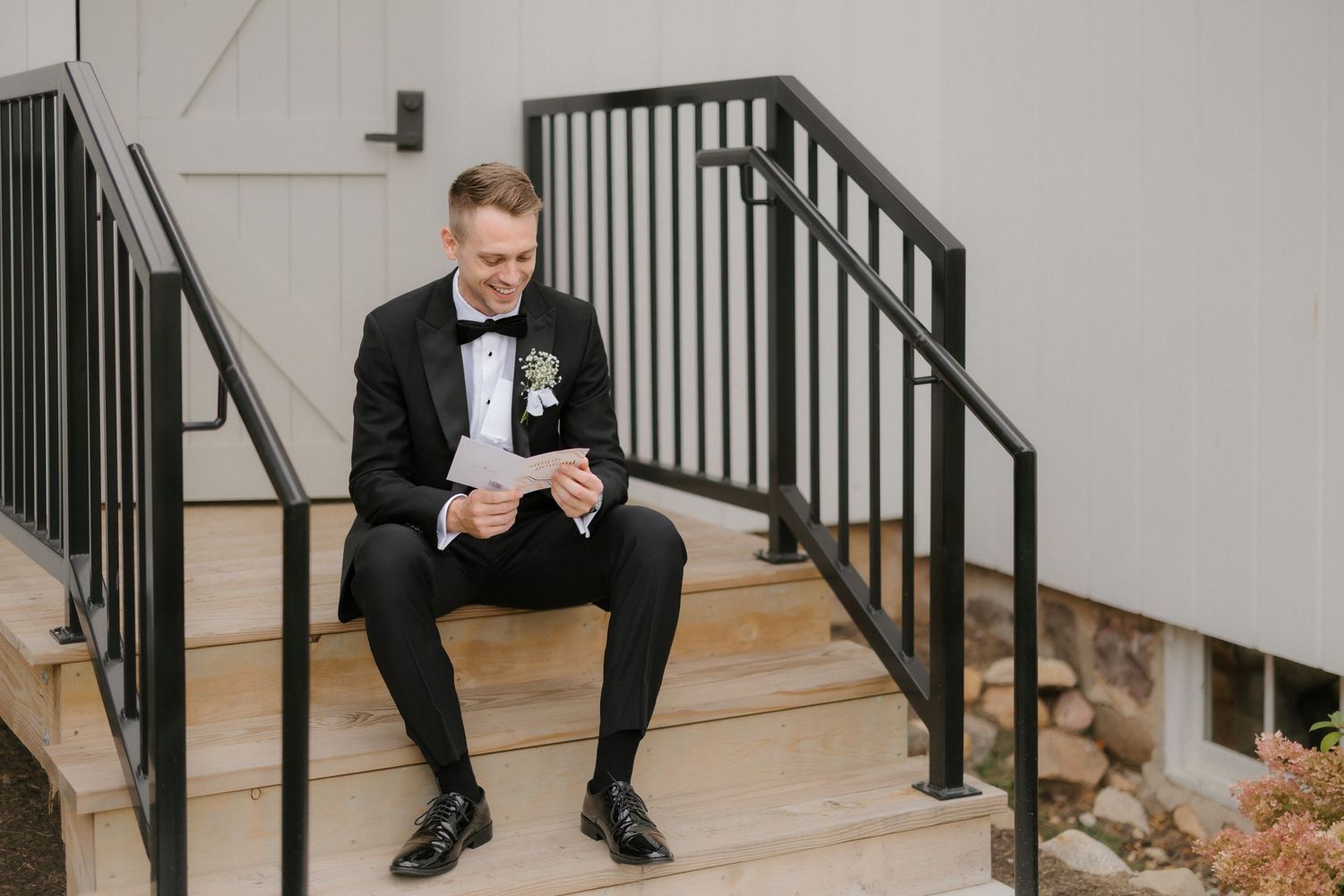 Smiling groom in black tuxedo reading letter while sitting on outdoor staircase steps.