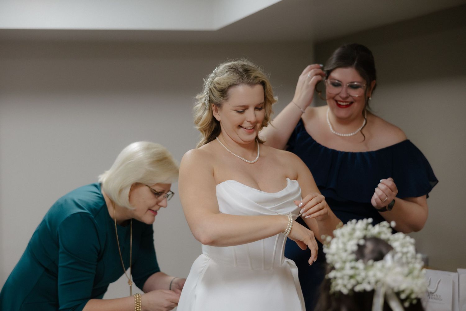 Bride in white gown smiles as two women in navy and teal help her prepare for her wedding ceremony.