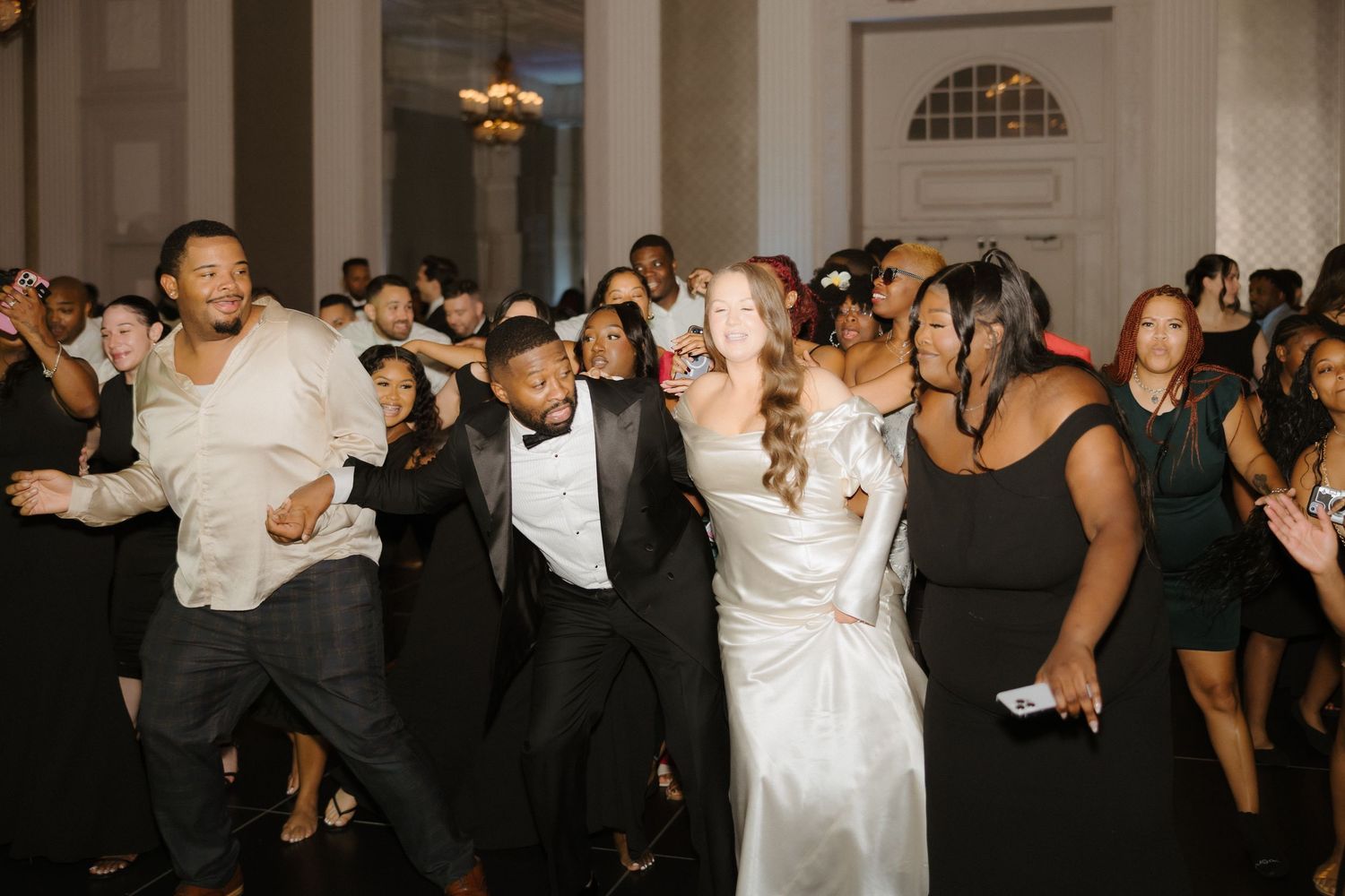 Wedding guests and couple dancing joyfully at elegant reception, bride in white gown, groom in tuxedo.
