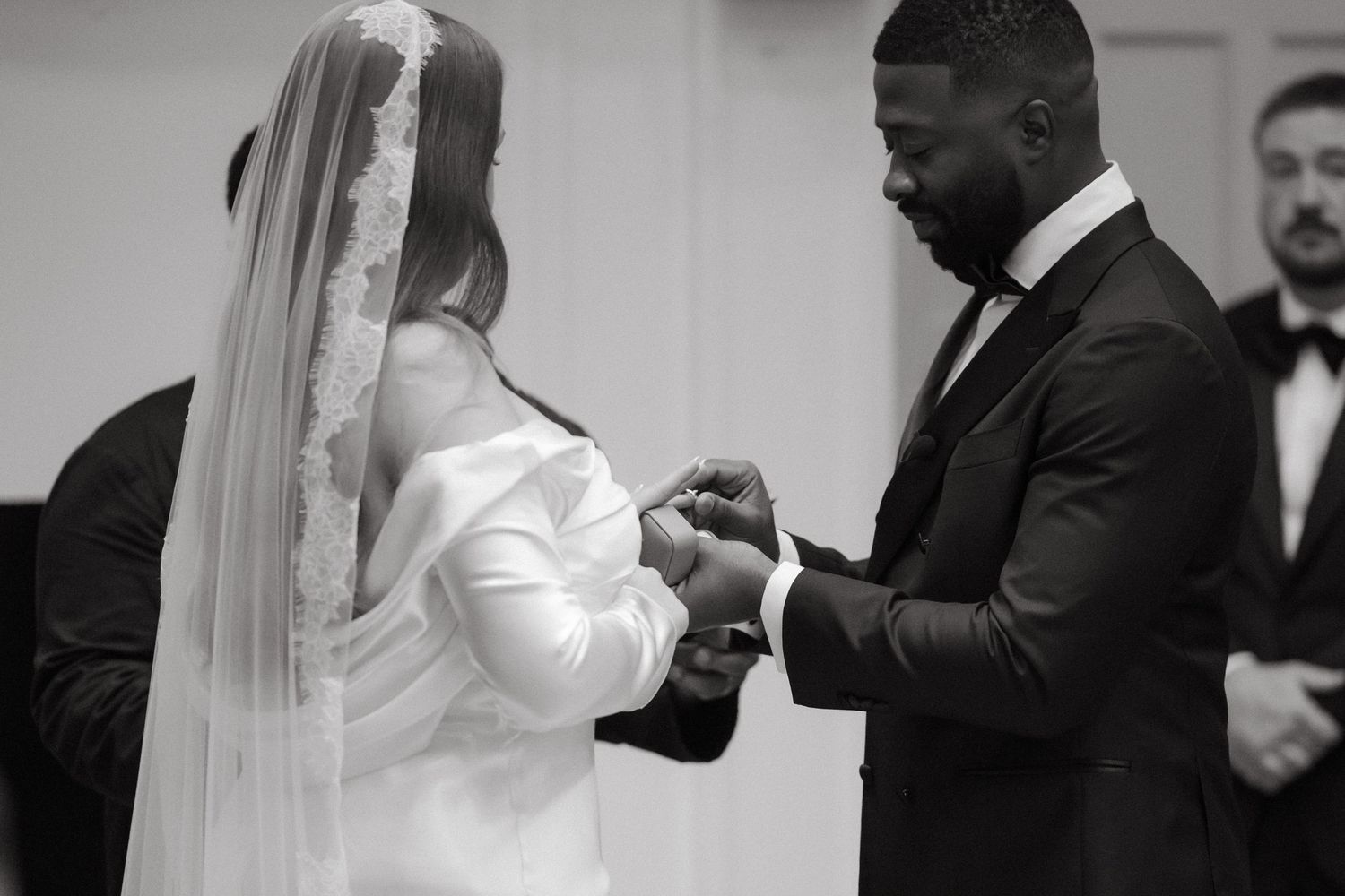 Black and white wedding photo of groom placing ring on brides finger during ceremony.