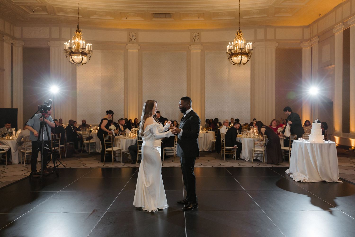 Couple sharing their first dance at an elegant wedding reception in a grand ballroom with chandeliers and seated guests.