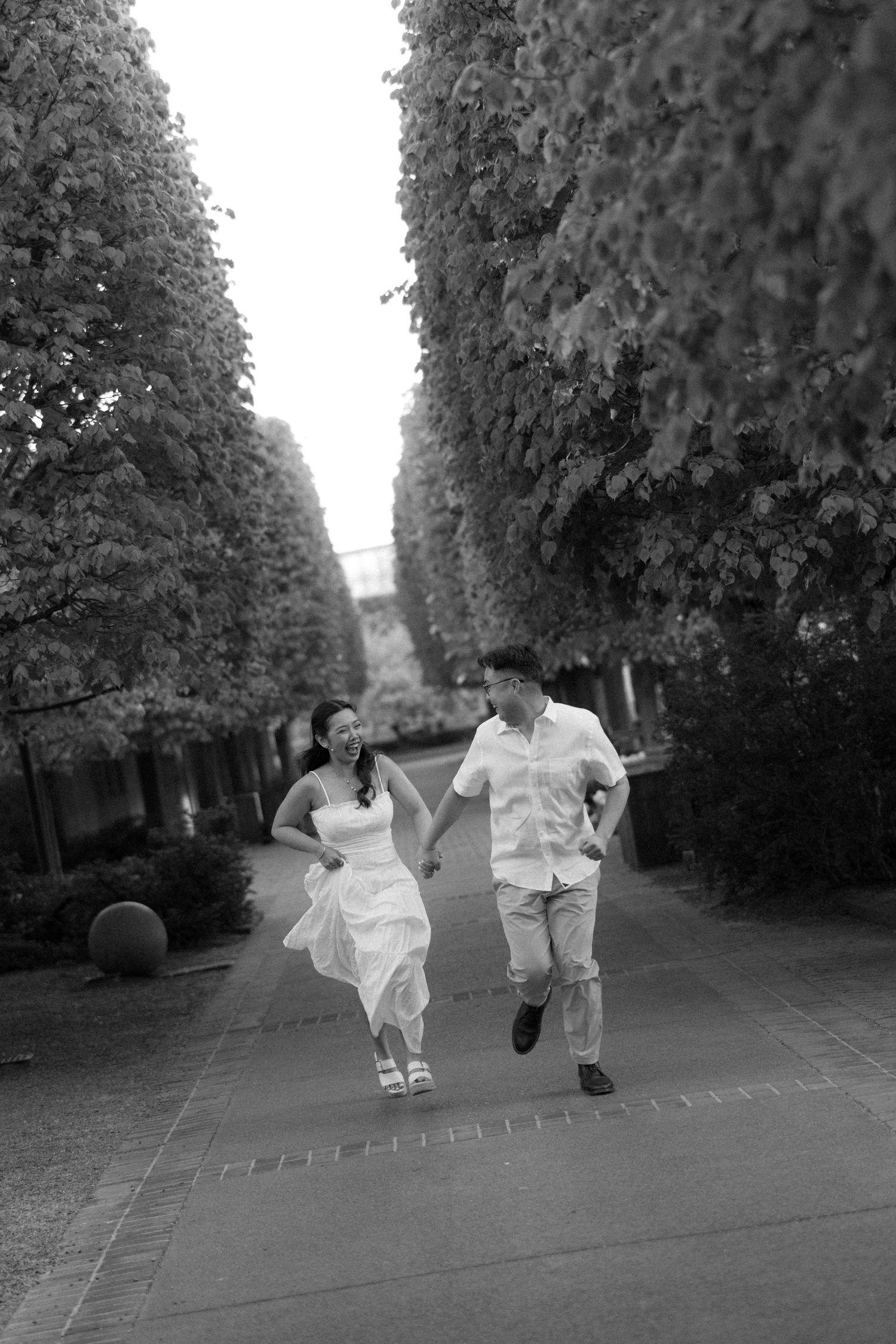Black and white photo of a couple holding hands and running joyfully down a tree-lined pathway.