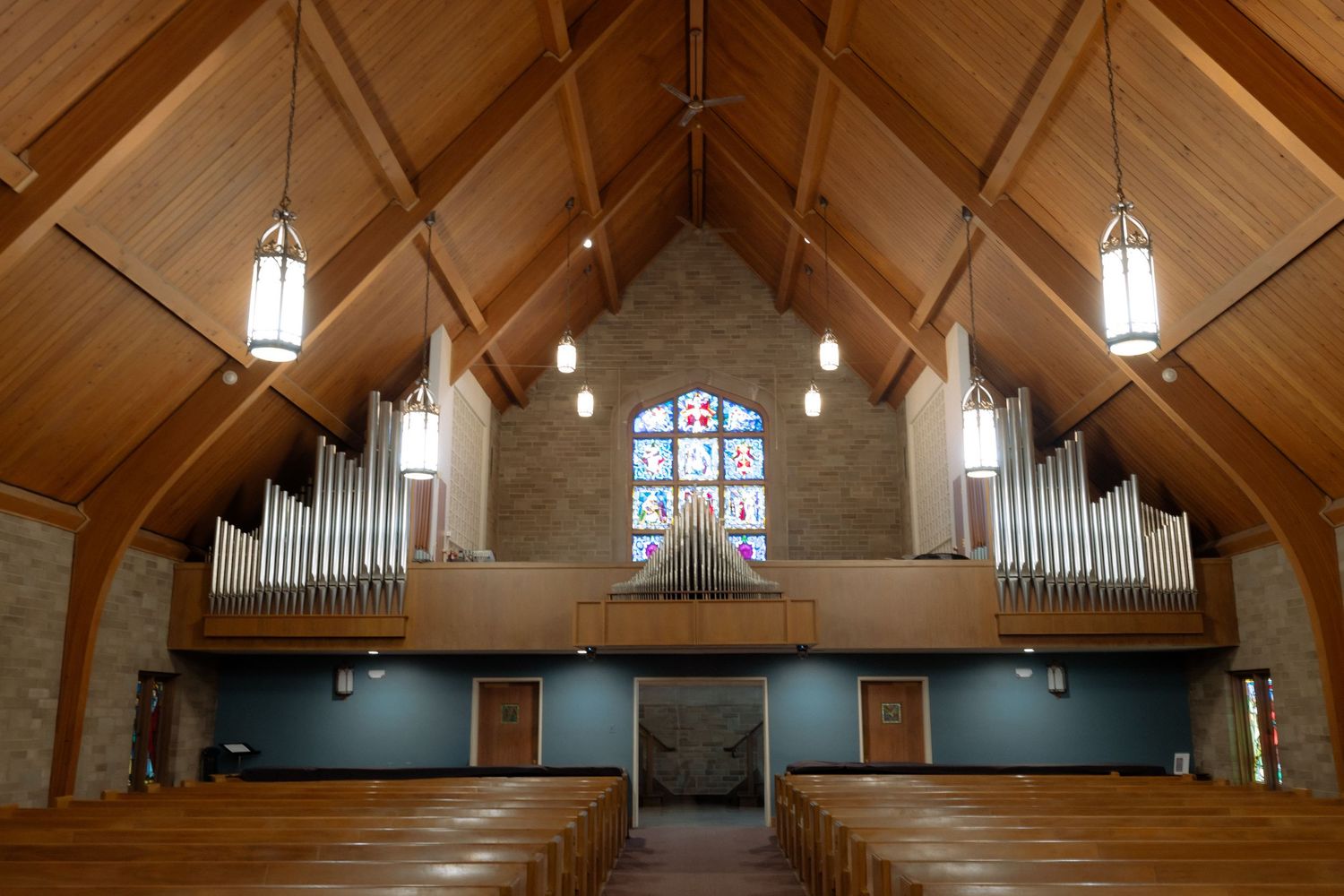 Church interior with wooden pews, pipe organ, stained glass window, and vaulted ceiling with pendant lights.