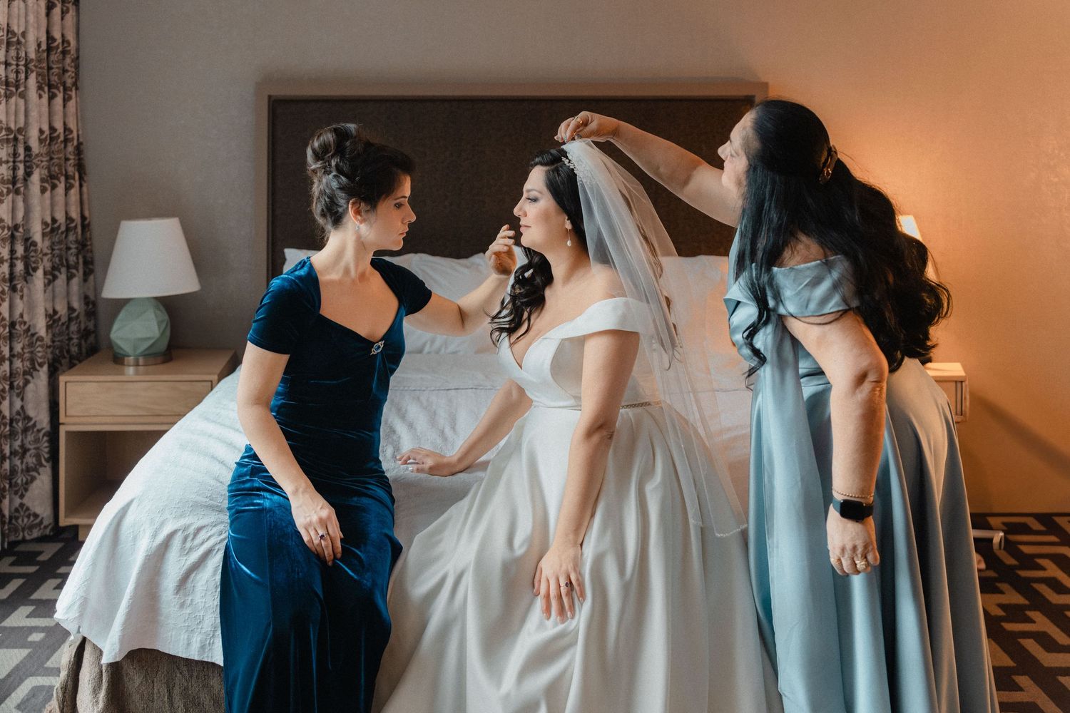 Bridesmaids in blue dresses help bride with veil and gown in a warmly lit hotel room.