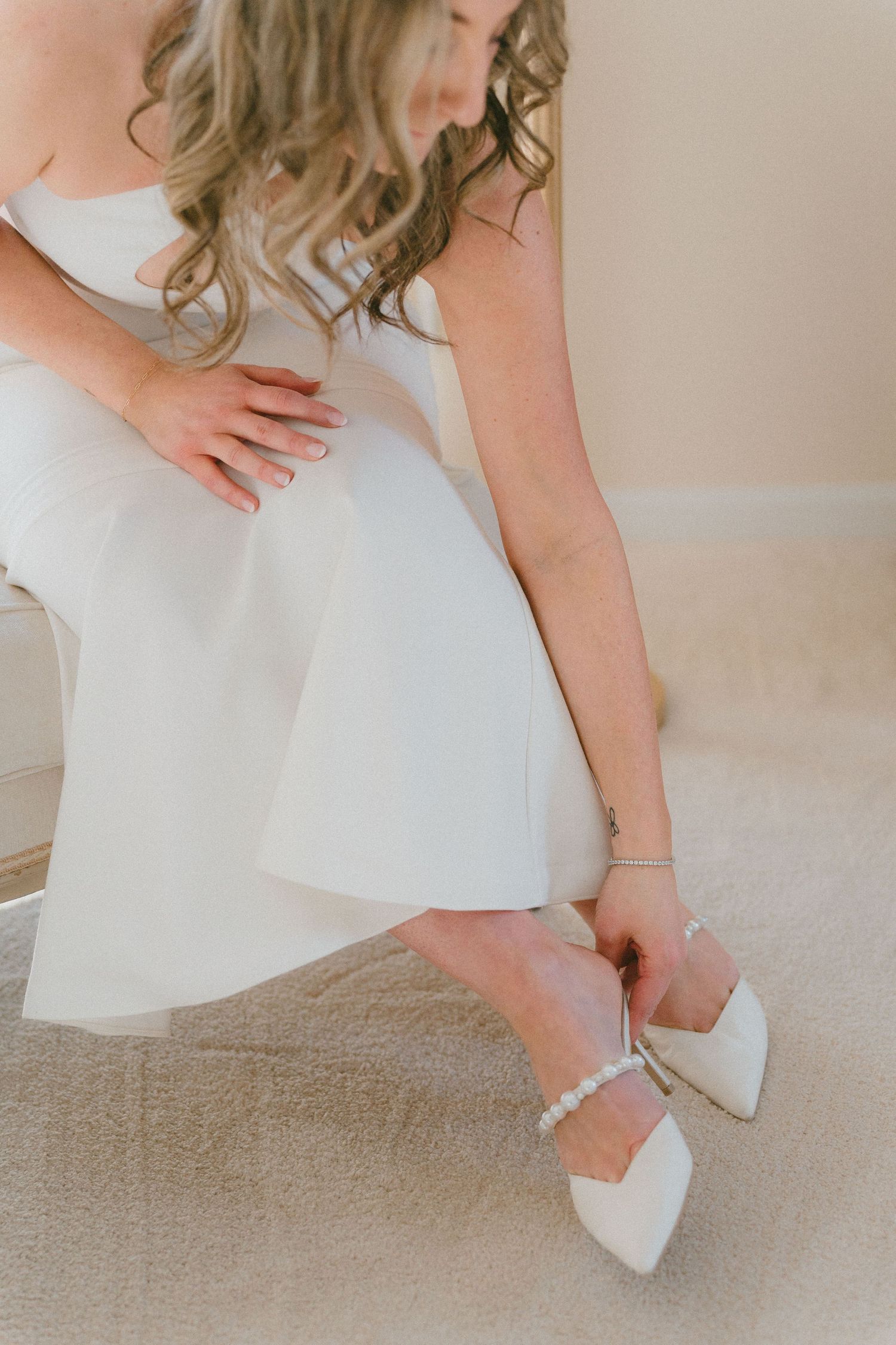 Bride in white dress fastening pearl ankle strap on pointed toe white wedding heels.