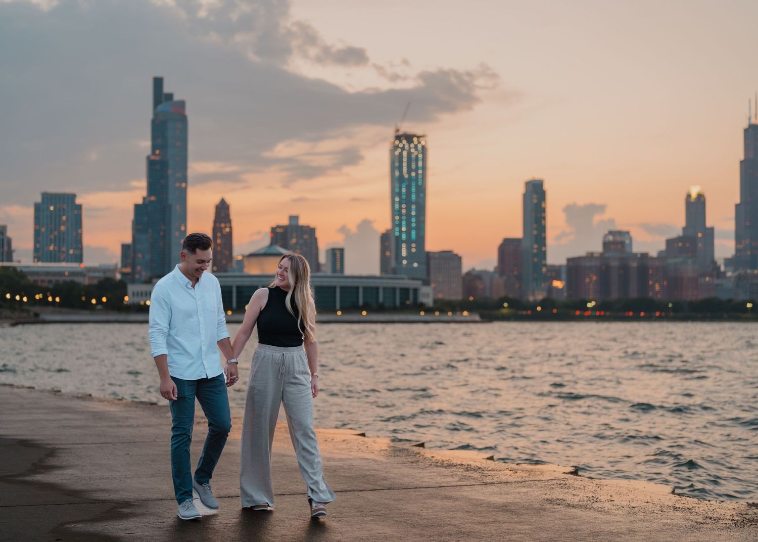 Couple holding hands walking along waterfront at sunset with Chicago city skyline in background.