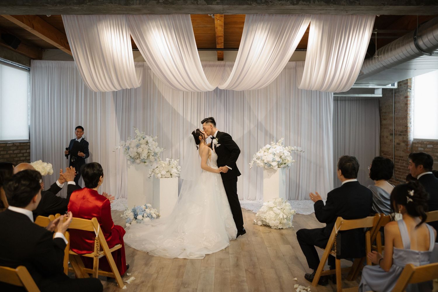 Bride and groom share first kiss at elegant indoor wedding ceremony with white draping and floral arrangements.