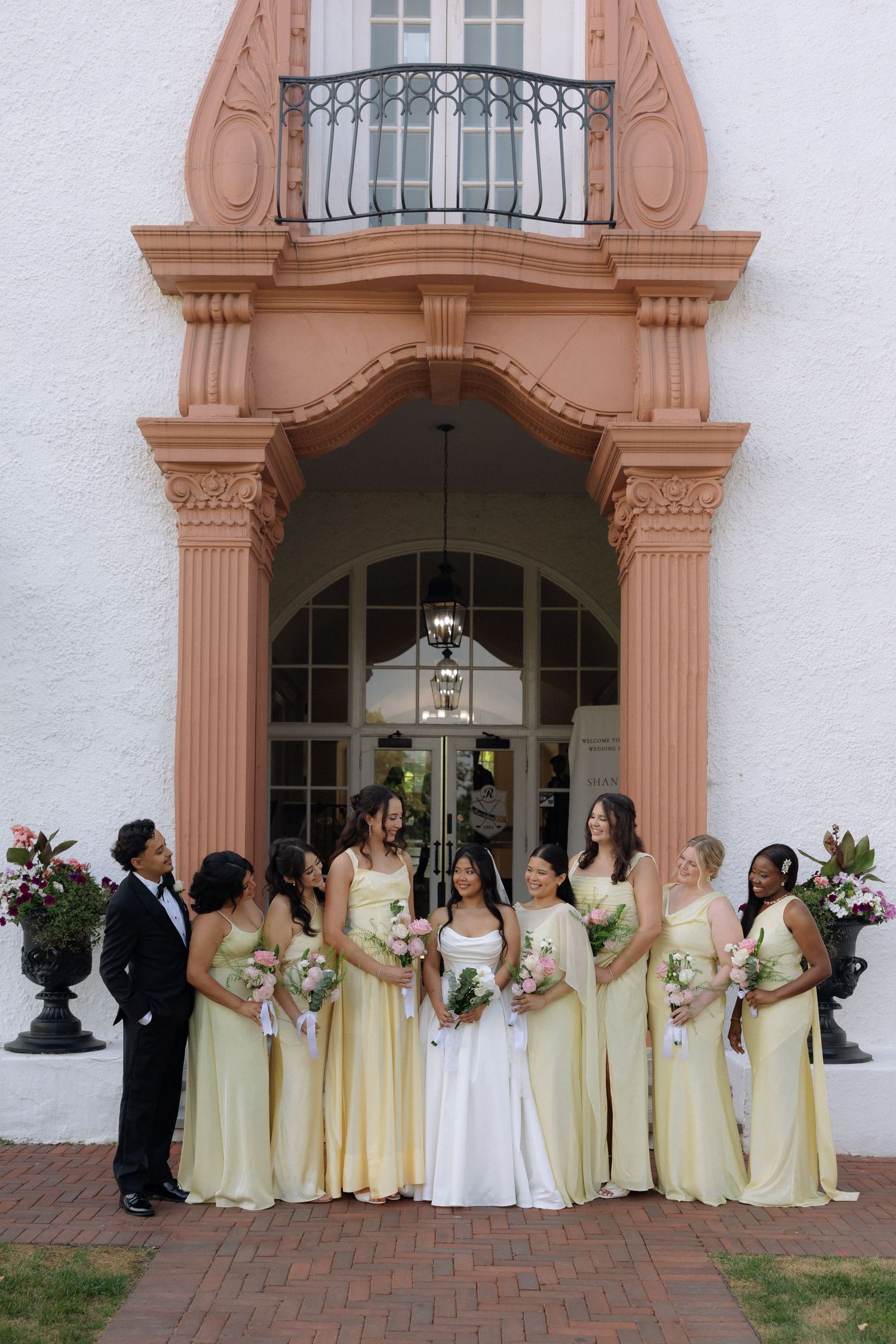 Bride in white gown poses with bridesmaids in yellow dresses outside elegant terracotta building.
