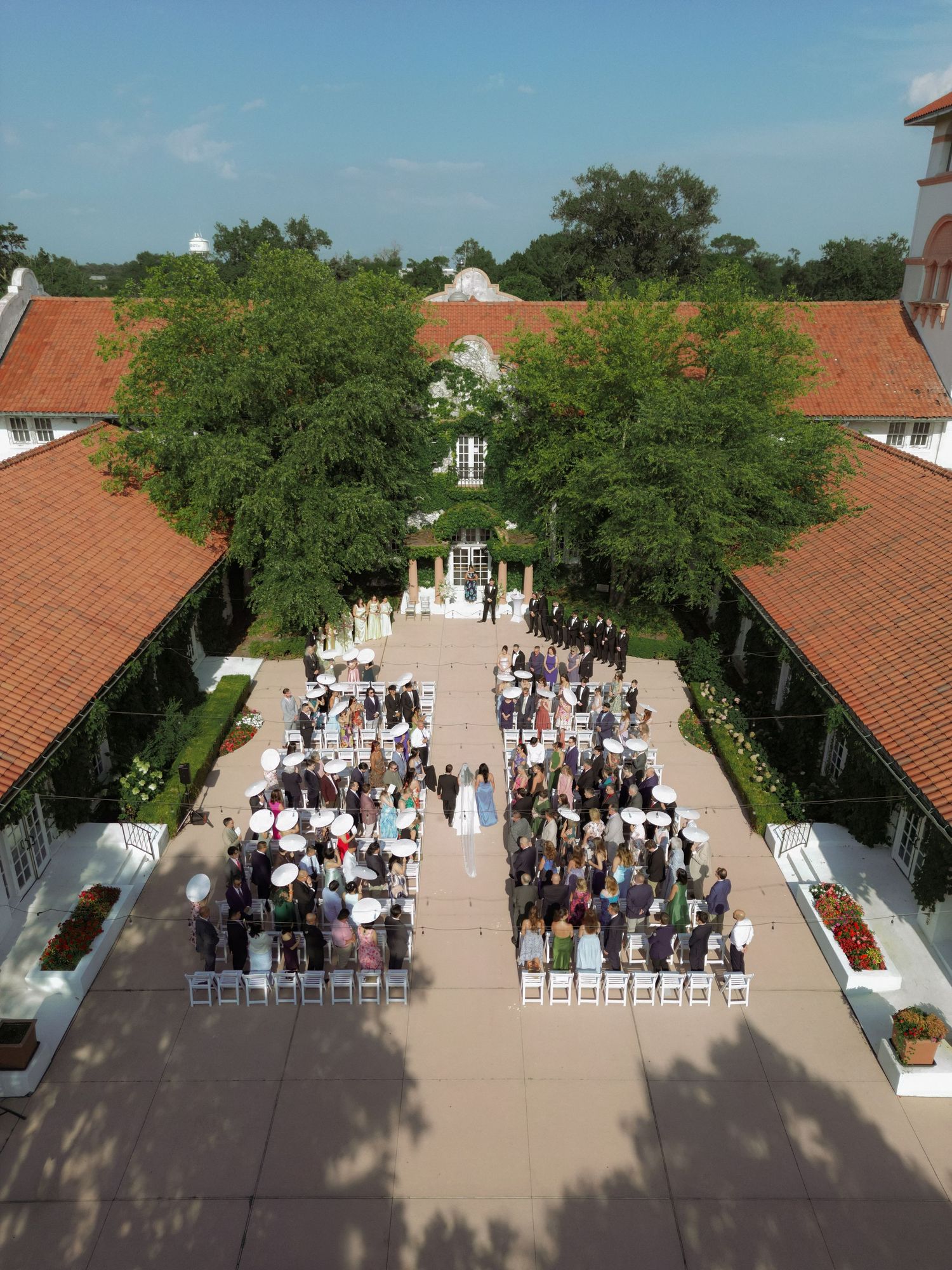Aerial view of an elegant outdoor wedding ceremony in a courtyard with red-tile roofed buildings and lush trees.