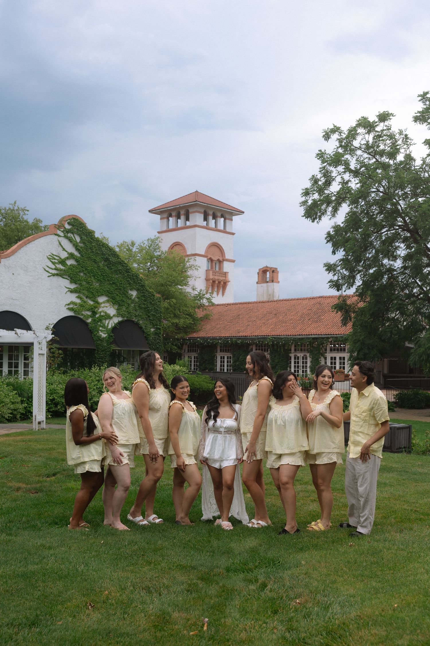 Bridal party in neutral outfits posing on green lawn before elegant building with red tile roof.