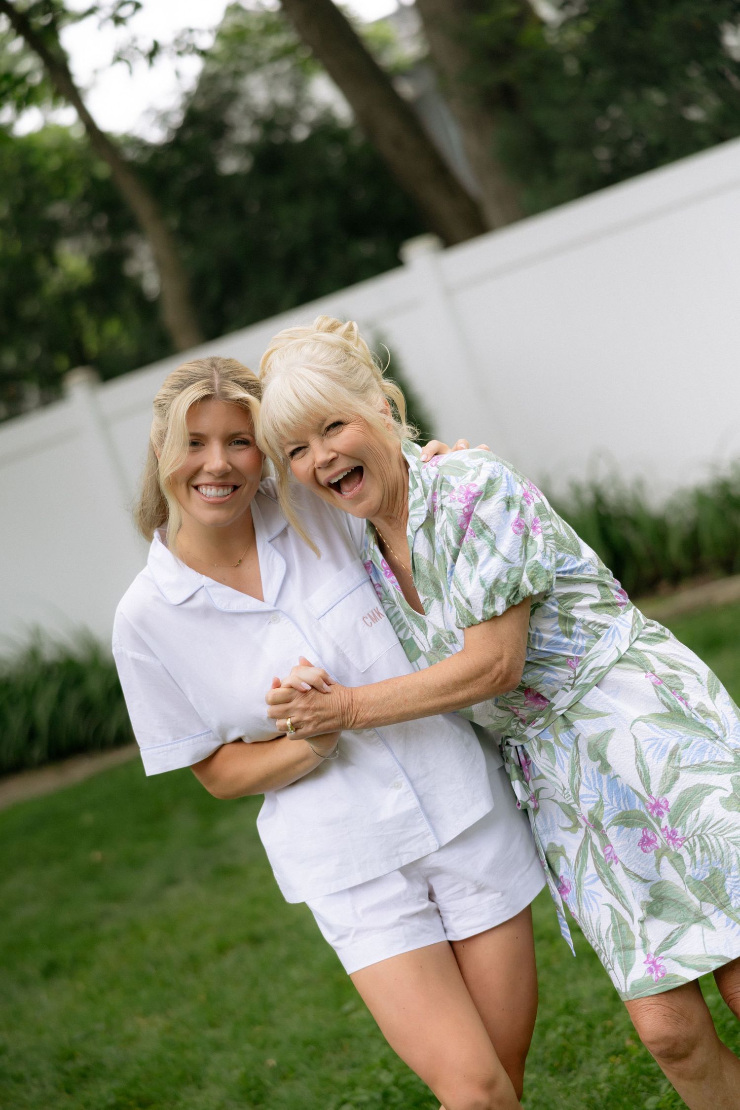 Two blonde women smiling and embracing joyfully in a lush green backyard setting.