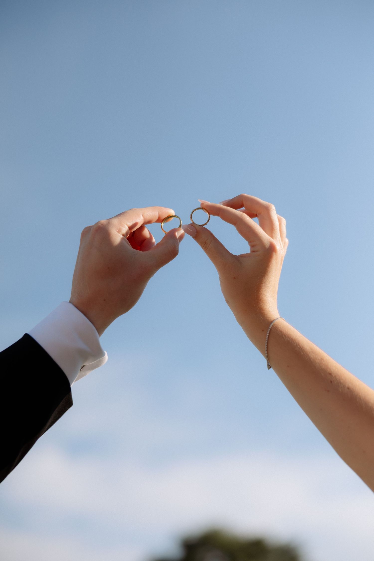 Bride and groom holding wedding rings up against blue sky, symbolizing their marriage commitment.