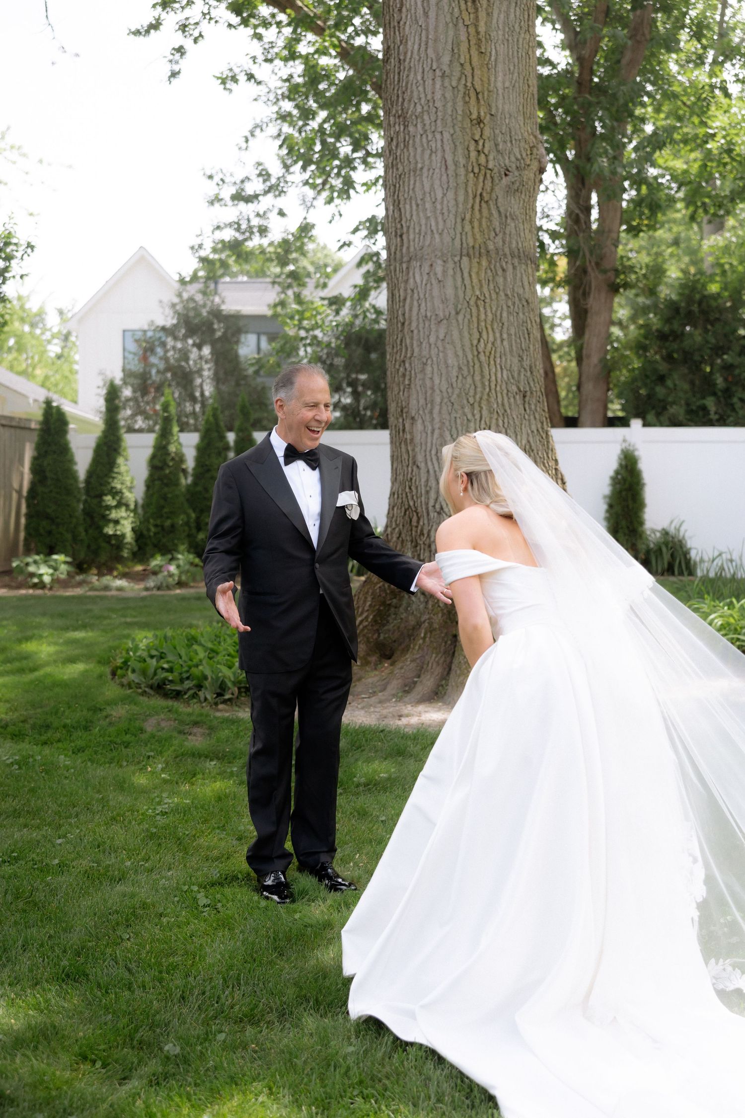 Father turns to see bride in flowing white gown during heartfelt first look in a green backyard.