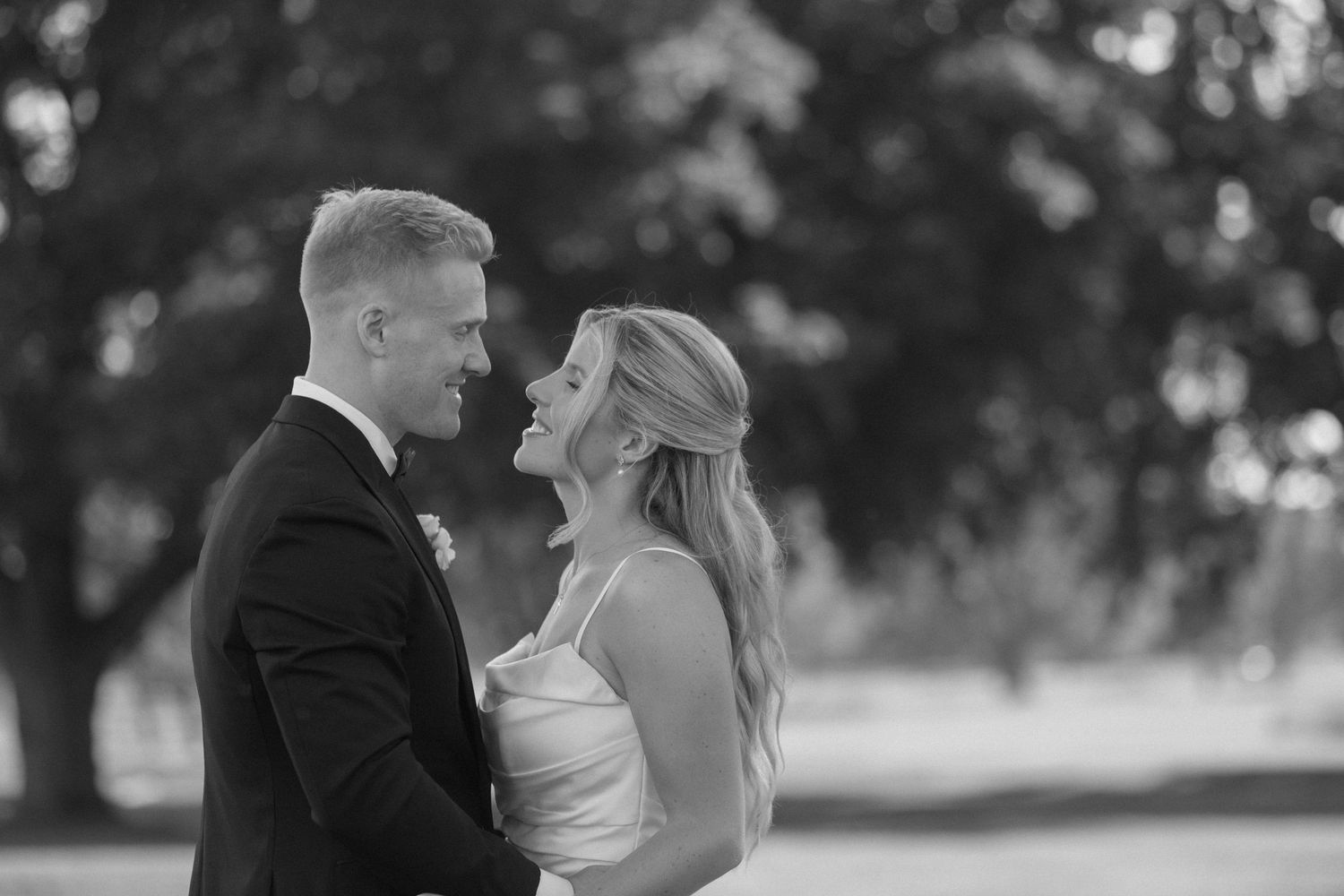 Black and white photo of bride and groom gazing at each other lovingly outdoors under trees.