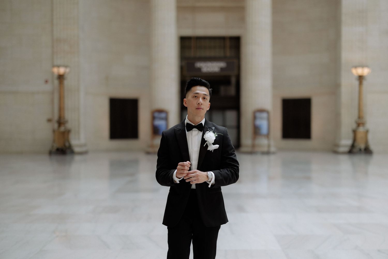 Groom in black tuxedo with bow tie stands in elegant marble hall, holding phone before wedding ceremony.