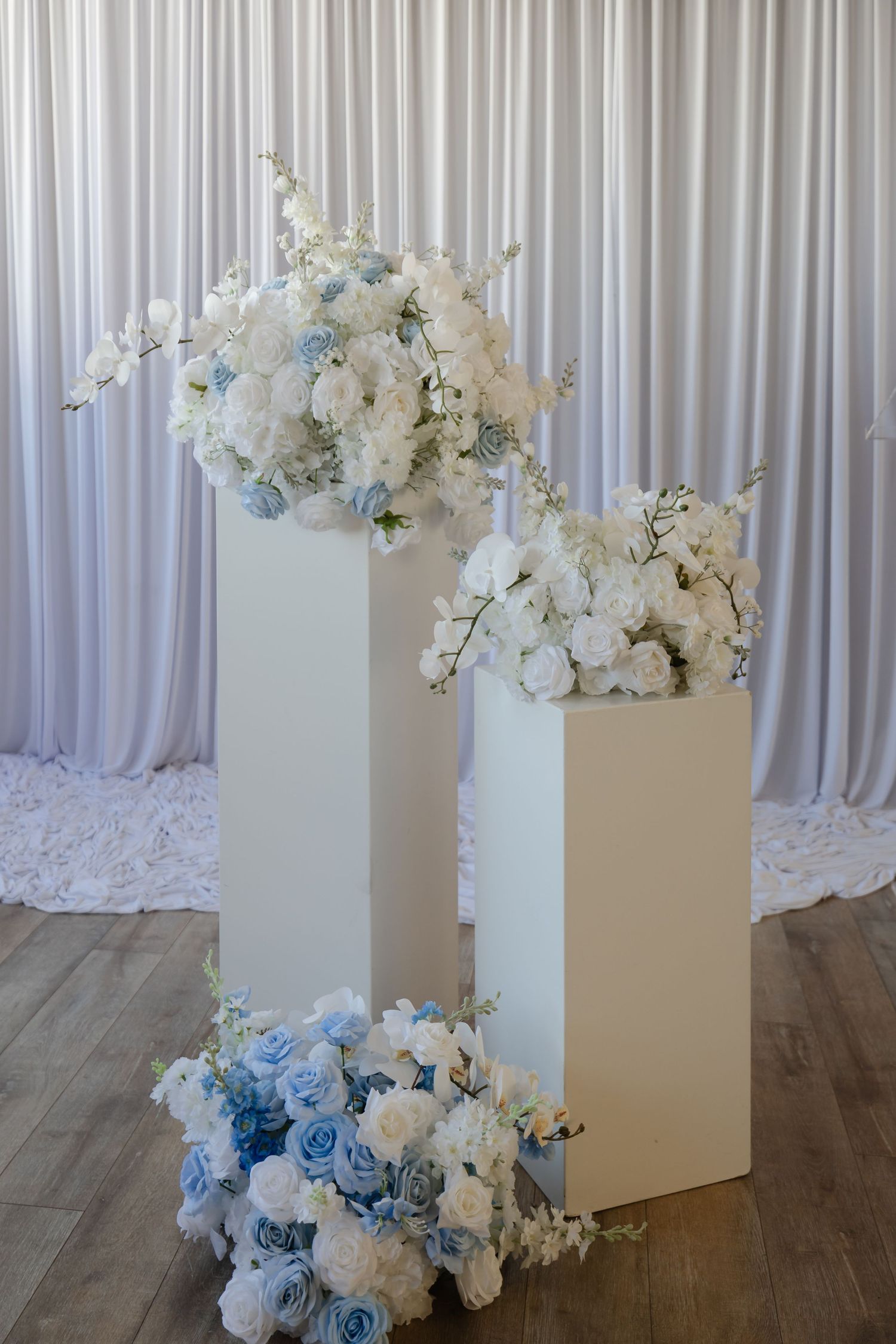Elegant blue and white rose bouquets on white pedestals against a white draped curtain wedding backdrop.