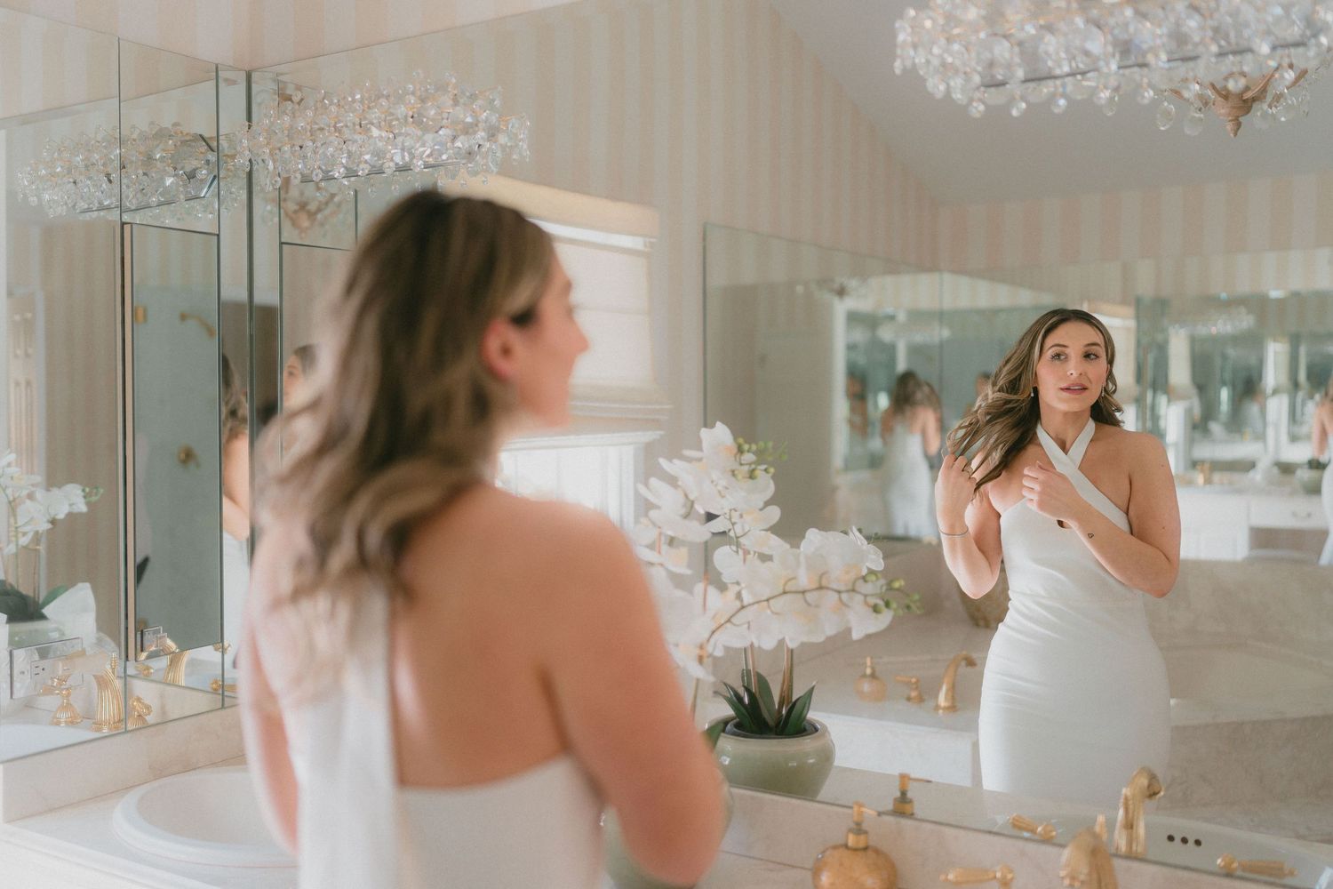 Bride in white halter dress admiring her reflection in elegant bathroom mirror.
