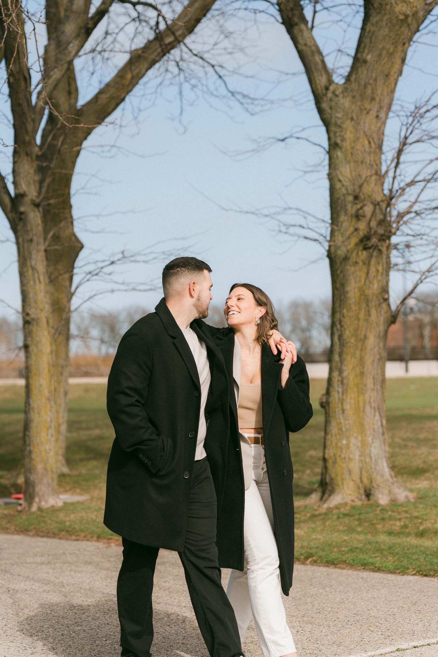 Happy couple in black coats sharing a romantic moment on a sunny park path with bare trees.