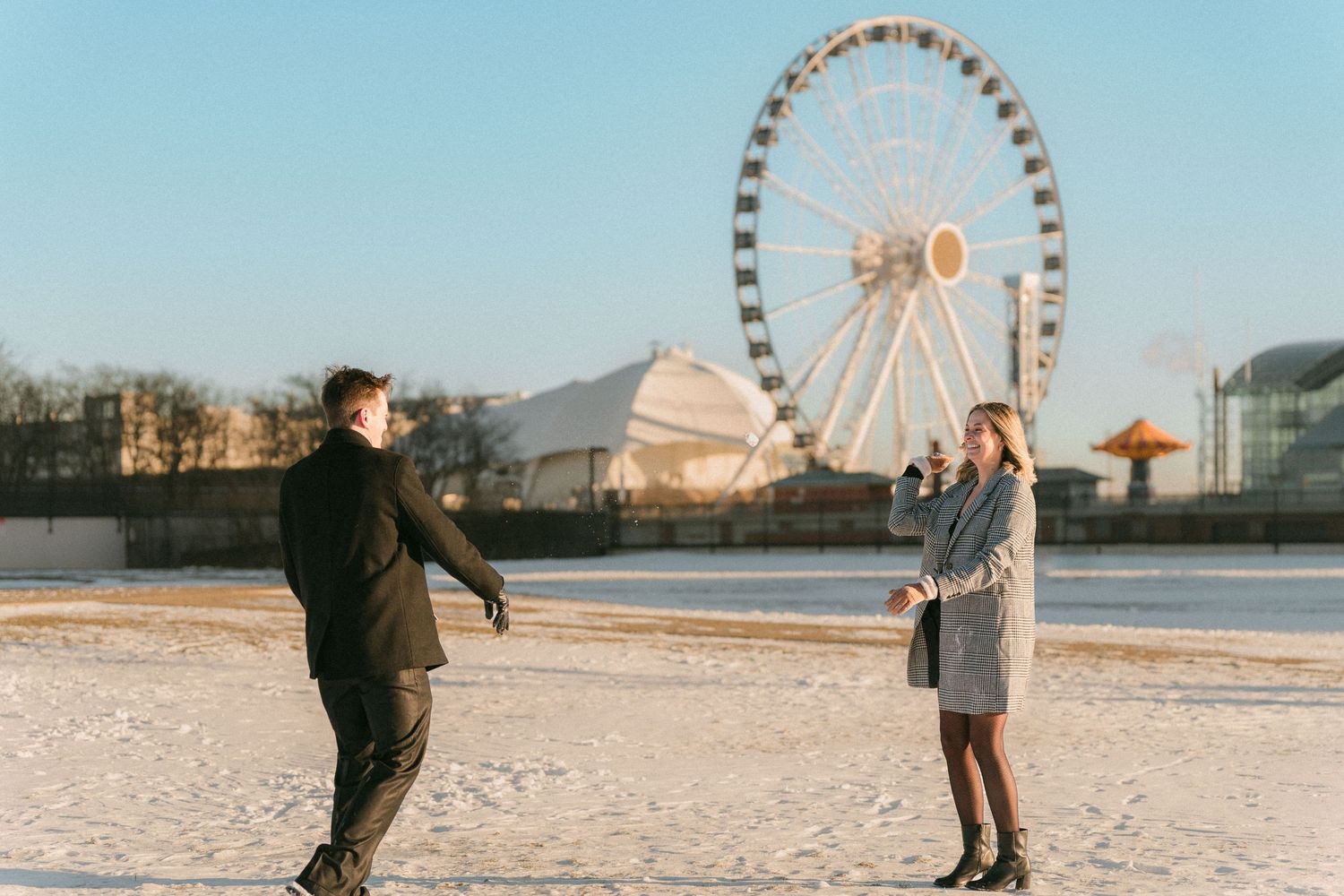 A playful couple faces each other near a Ferris wheel on a bright snowy winter day.