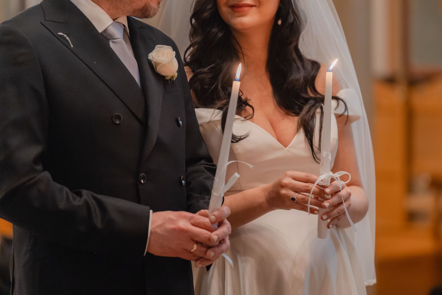 Close-up of bride and groom holding lit white unity candles together during elegant church wedding ceremony.