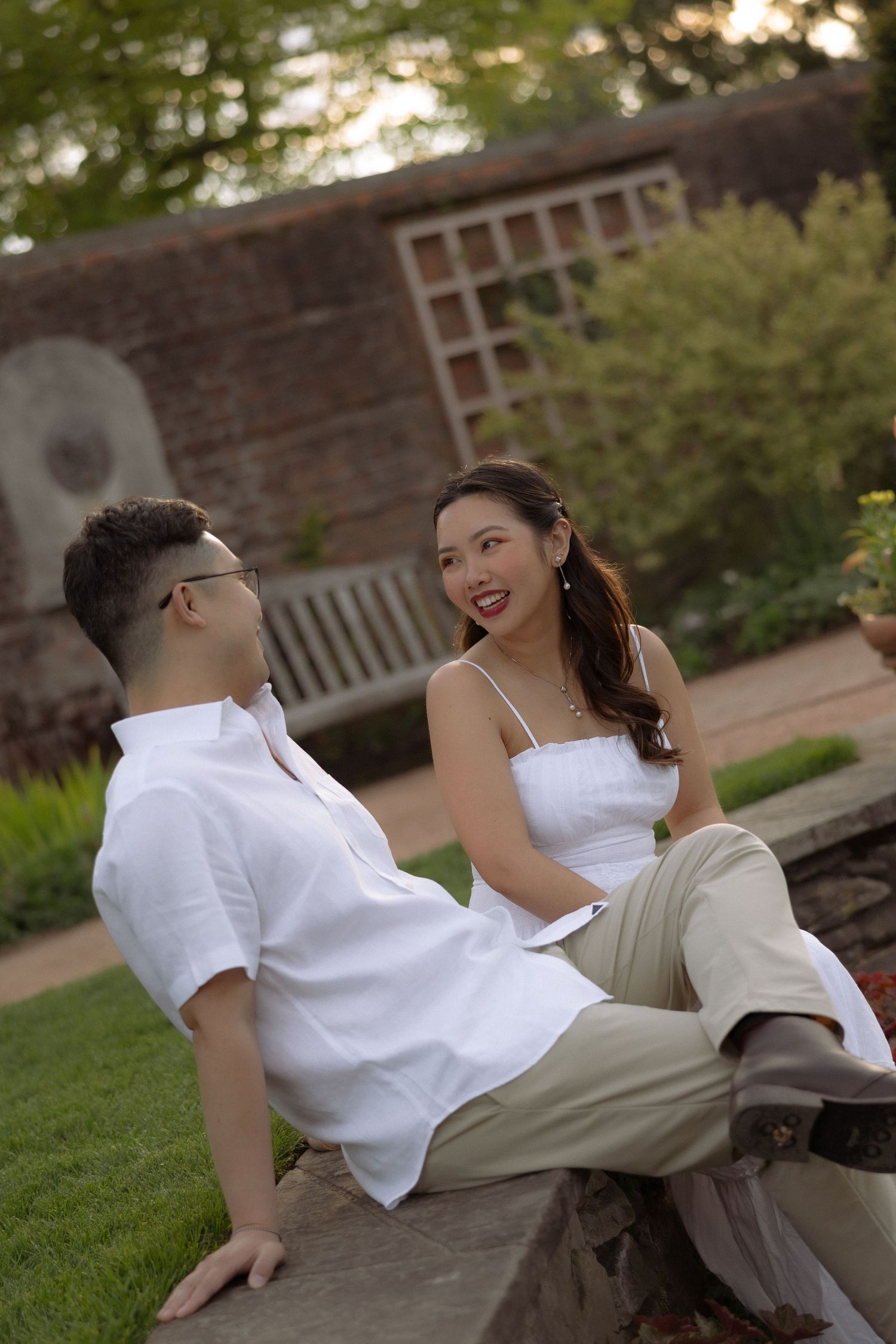 A young Asian couple in matching white outfits sit together on stone steps in a lush garden, smiling and conversing.