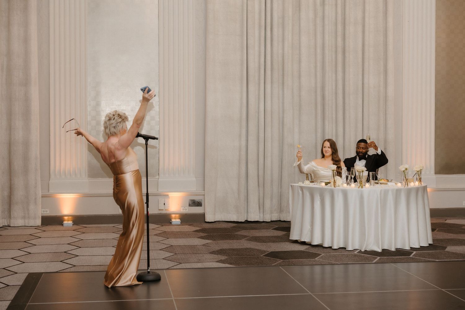 A performer in a gold gown sings at a microphone while a couple celebrates at a sweetheart table in an elegant ballroom.