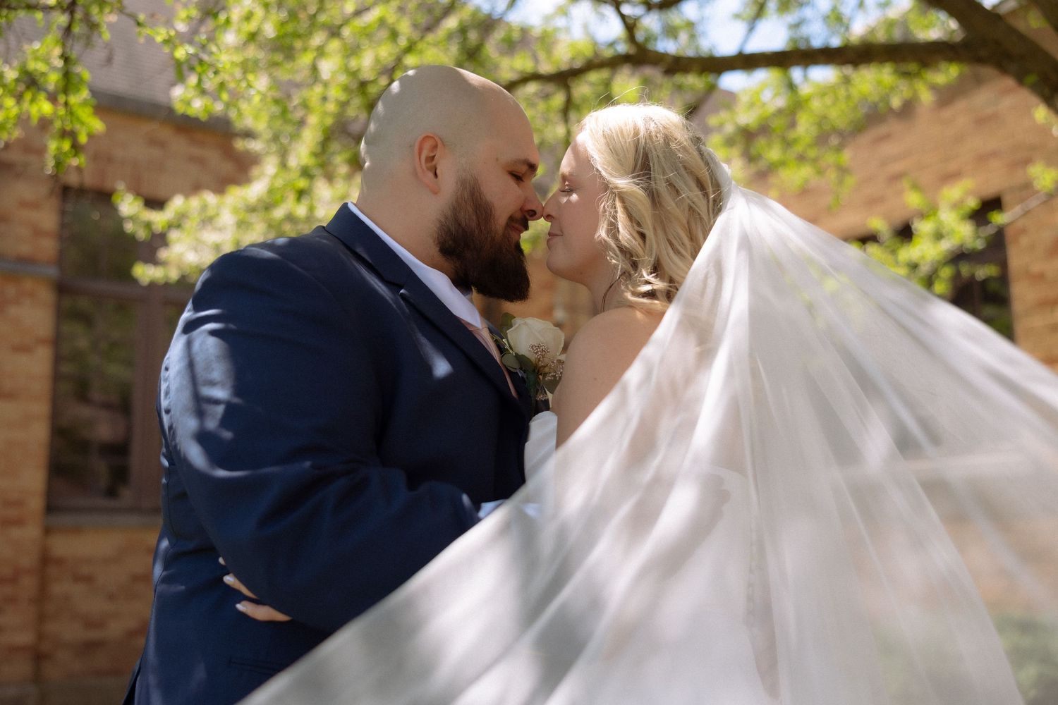 A bride and groom share a romantic kiss outdoors, with the bride's veil flowing in the breeze on their wedding day.