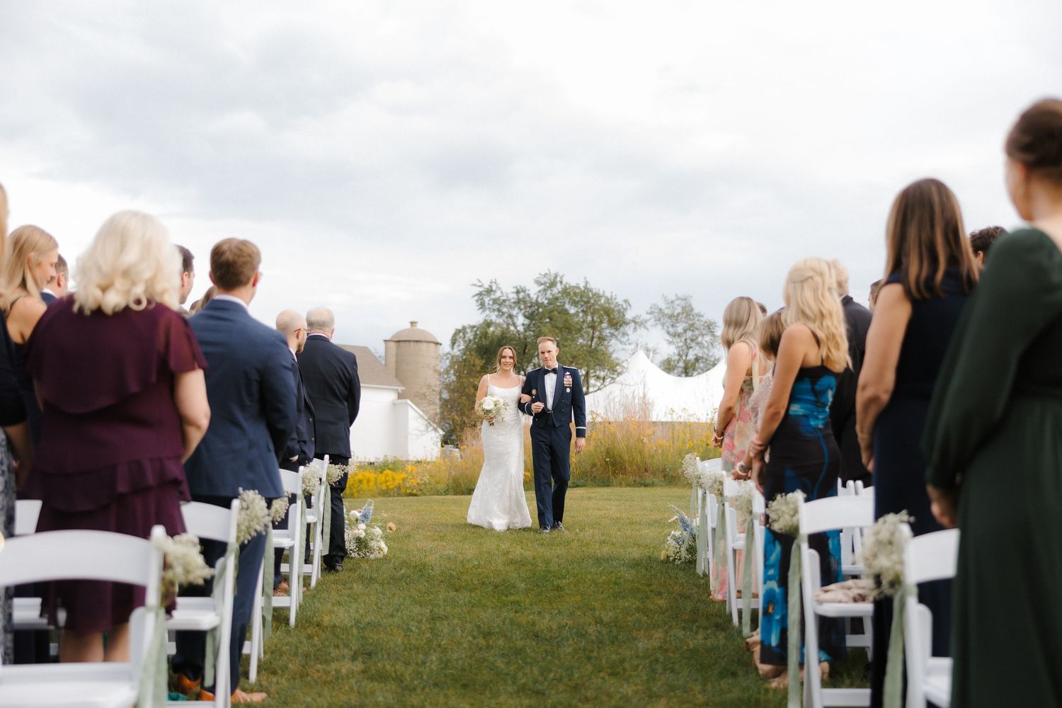 Bride and groom walk down outdoor aisle lined with white chairs and seated wedding guests on cloudy day.