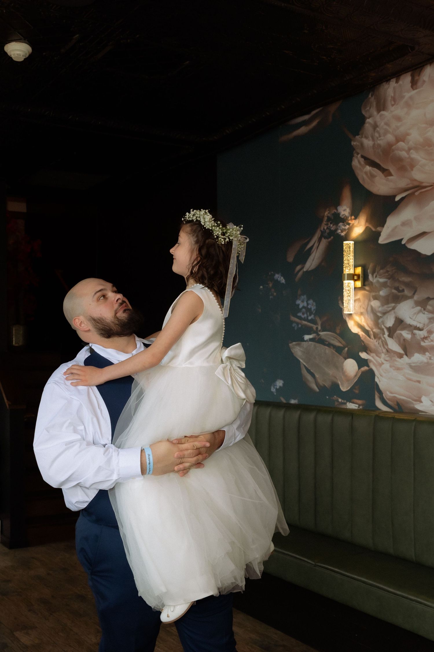 Bald groom in white shirt lifts flower girl in white tulle dress and floral crown at elegant wedding venue.