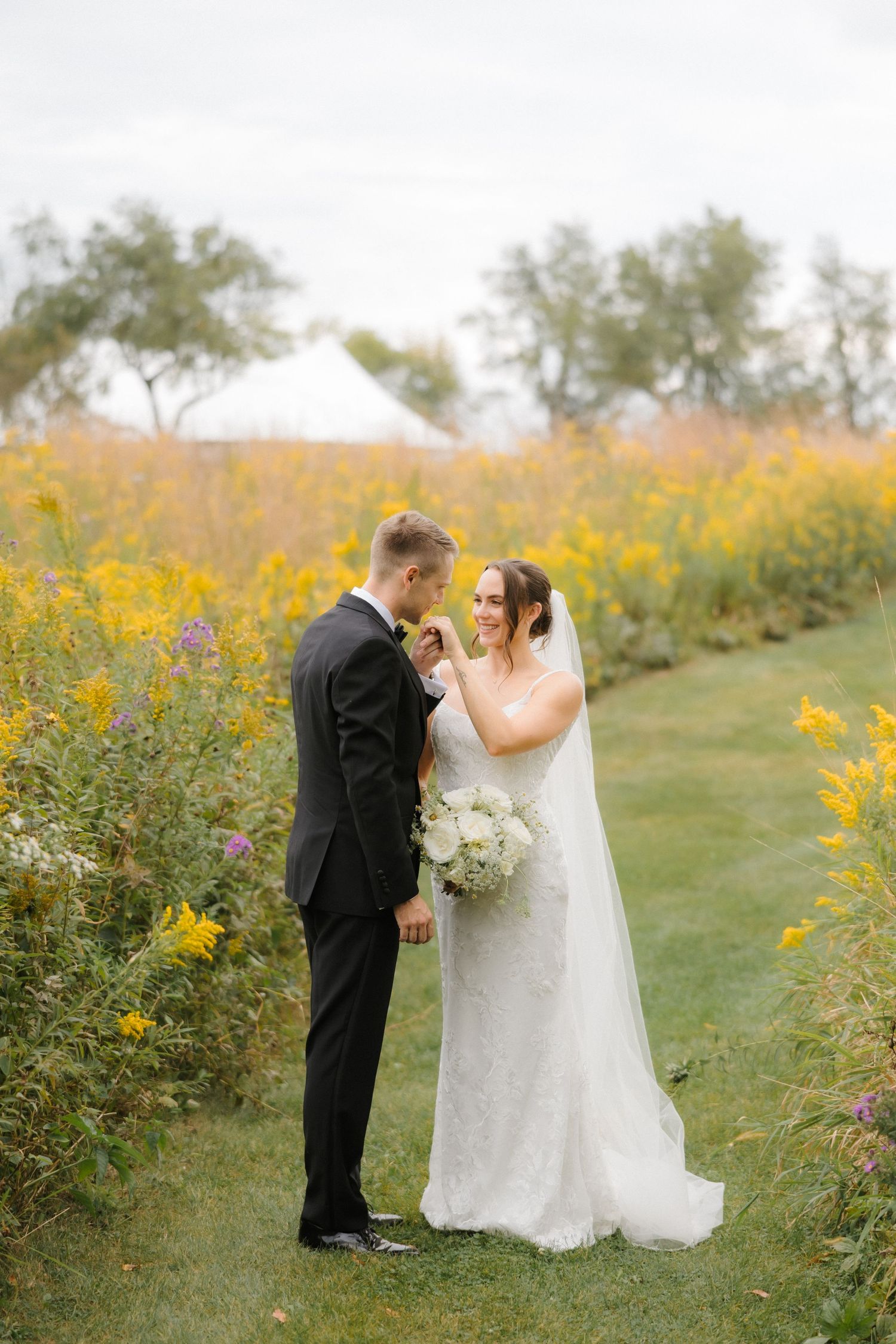 Groom kissing bride's hand as she smiles holding a white bouquet in a field of yellow wildflowers.