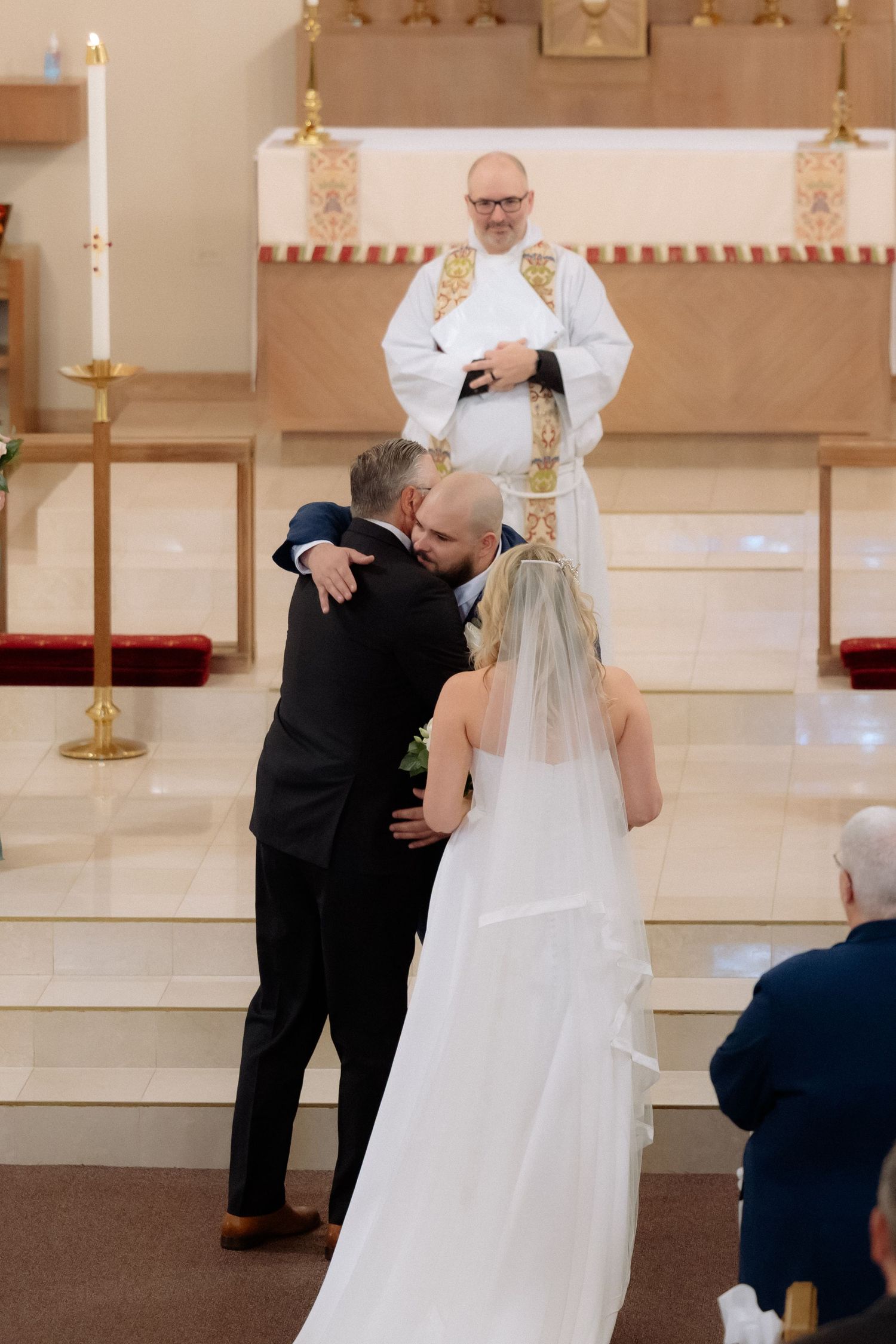 Groom embracing father of the bride at church altar during wedding ceremony with priest officiating.
