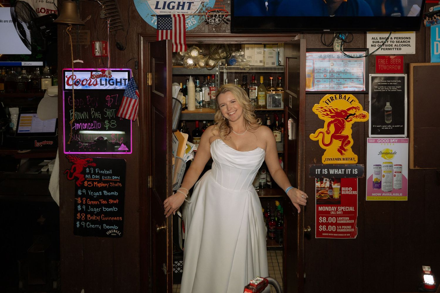 Blonde bride in white wedding dress posing at a bar entrance with neon beer signs.