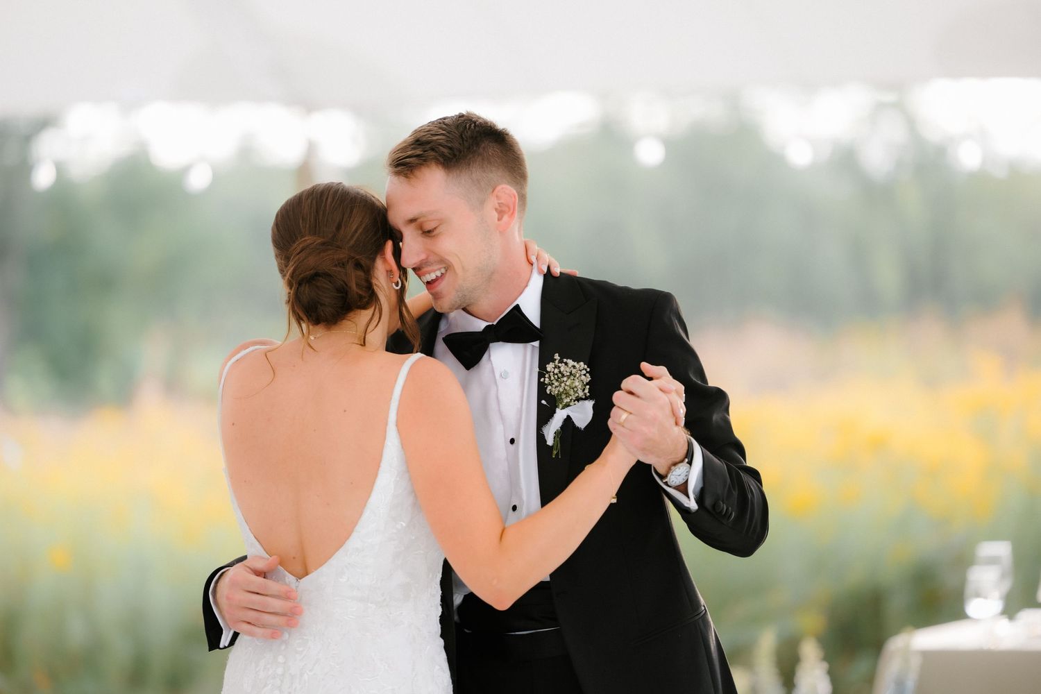 Bride and groom share a romantic first dance outdoors under a white tent with colorful wildflower fields behind them.