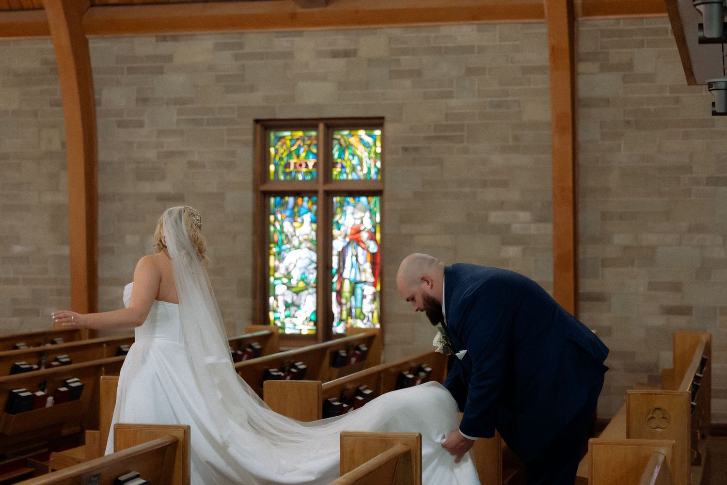 Bride in white gown and veil walks down church aisle with stained glass windows while groom adjusts her train.