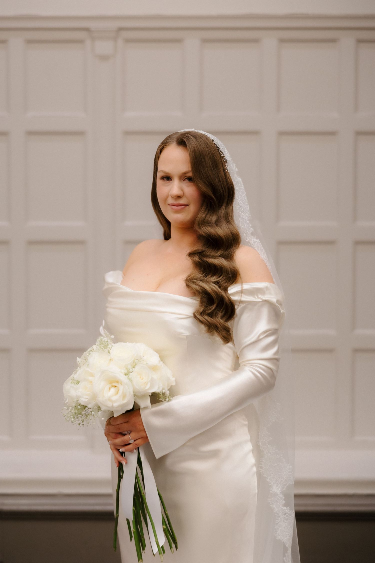Elegant bride in off-shoulder white satin gown with veil holds white peony bouquet, posing before white paneled wall.