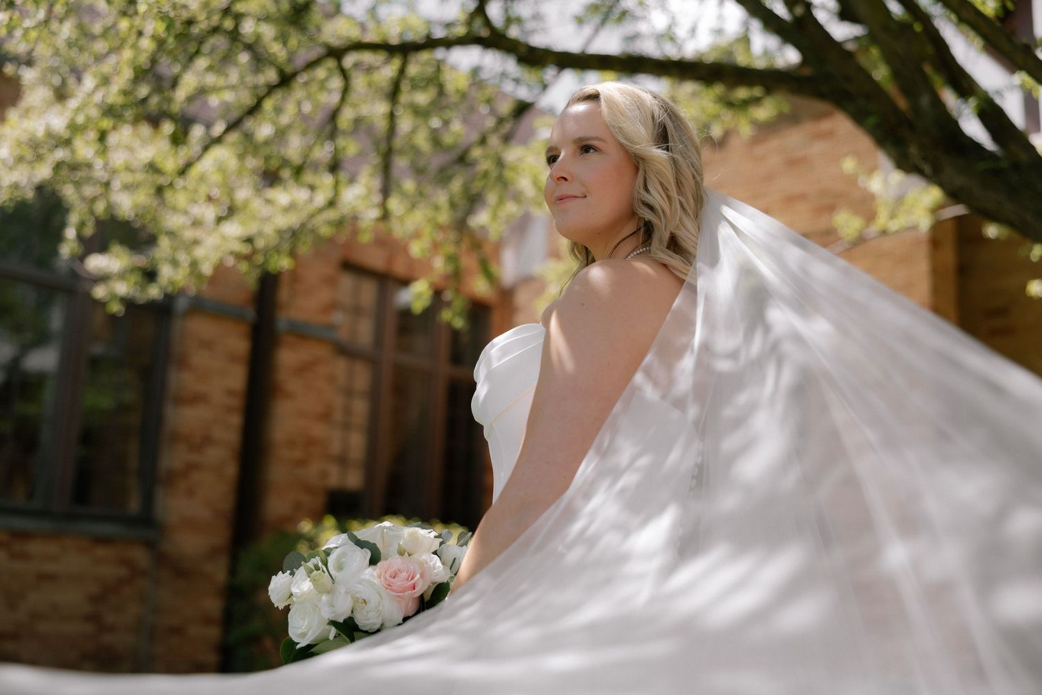 Bride in white gown and flowing veil holds bouquet of white peonies outdoors under blooming spring trees.