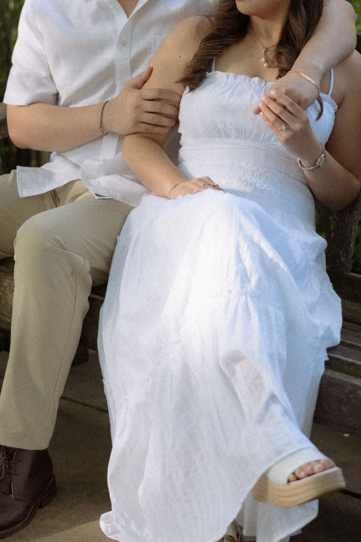 Couple sitting together on a bench, woman in white strapless dress, man in white shirt and khaki pants, holding hands.