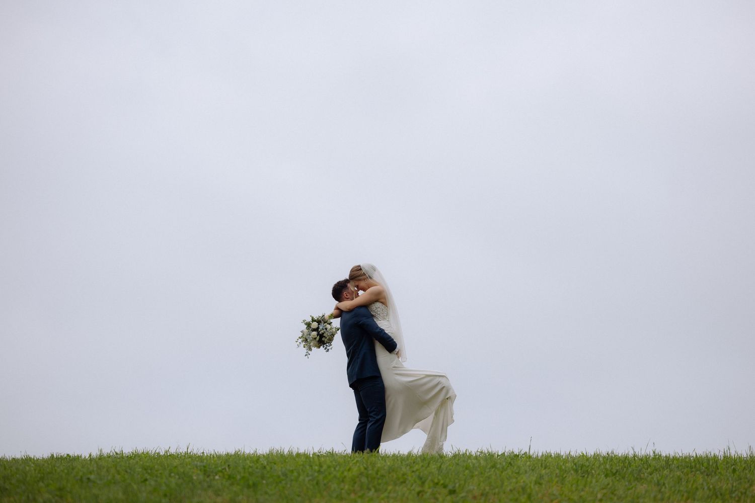 Groom lifts bride on a green hillside against a gray sky in a romantic wedding portrait.