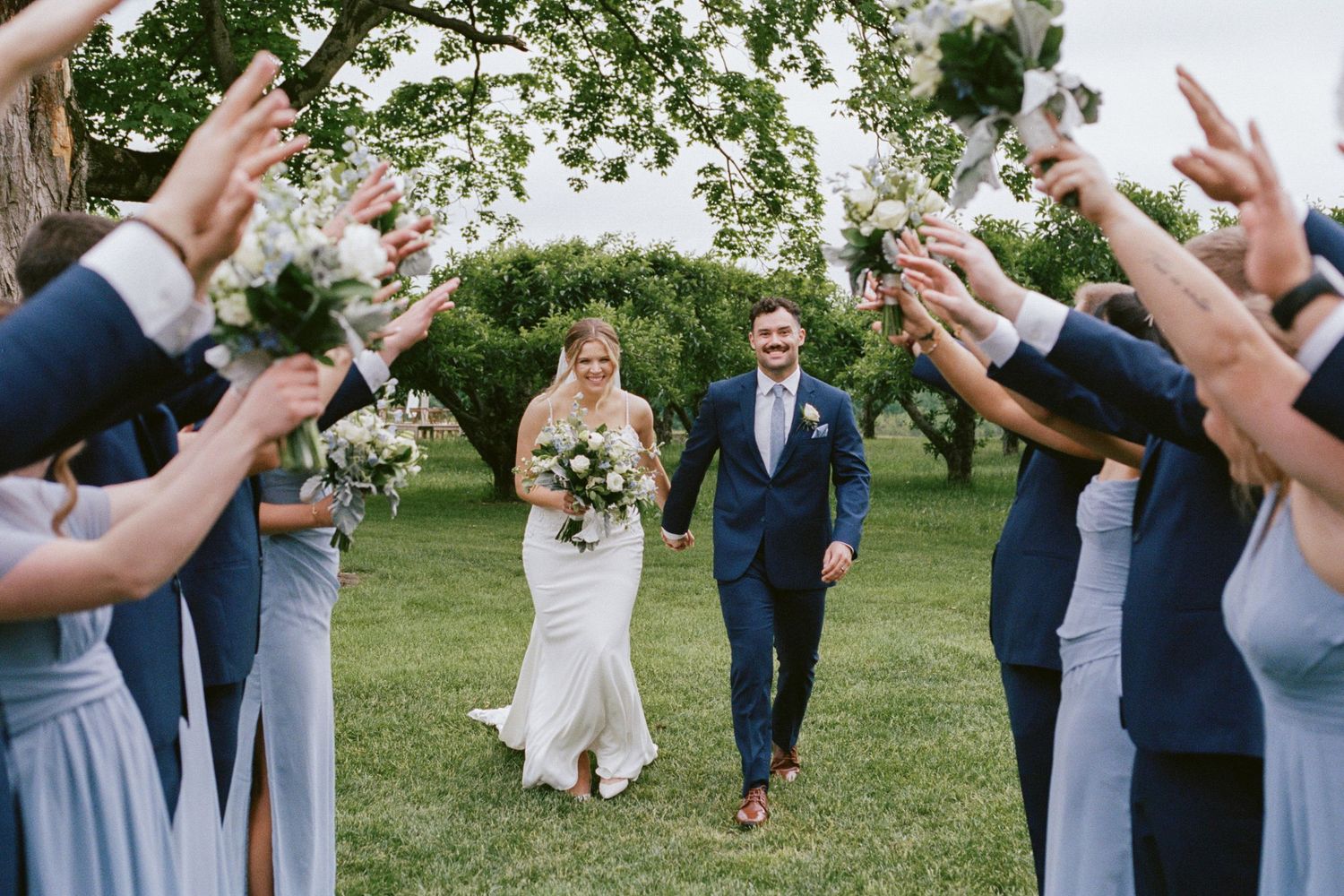 Bride and groom walk through a floral tunnel of cheering wedding guests outdoors at a garden ceremony.