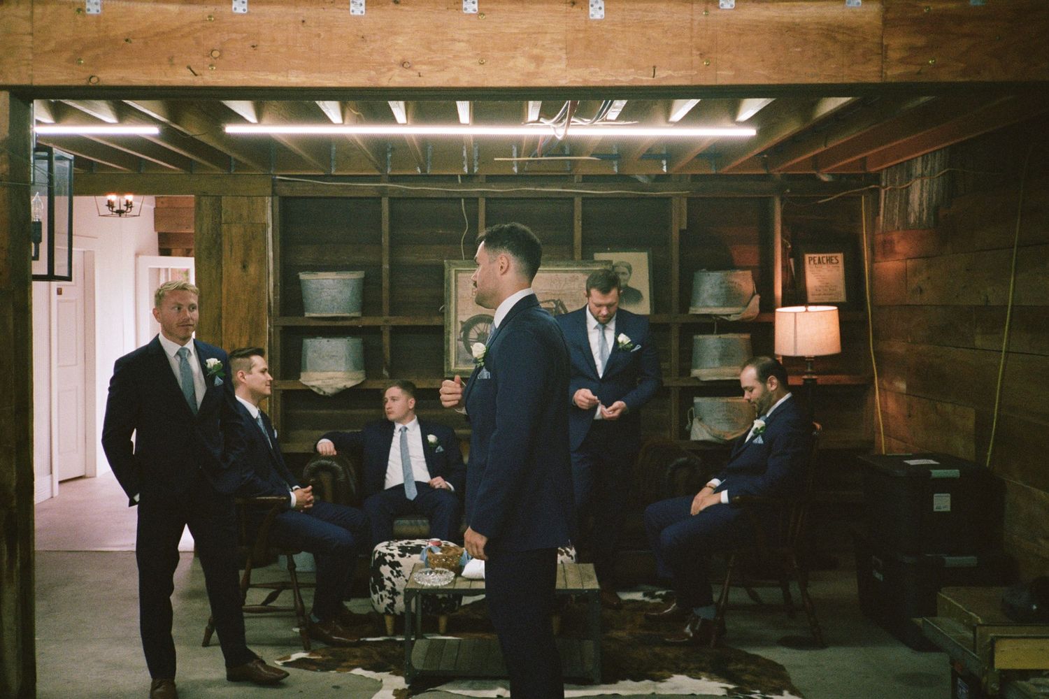 Groomsmen in navy suits gather in a rustic wood-paneled room before a wedding ceremony.