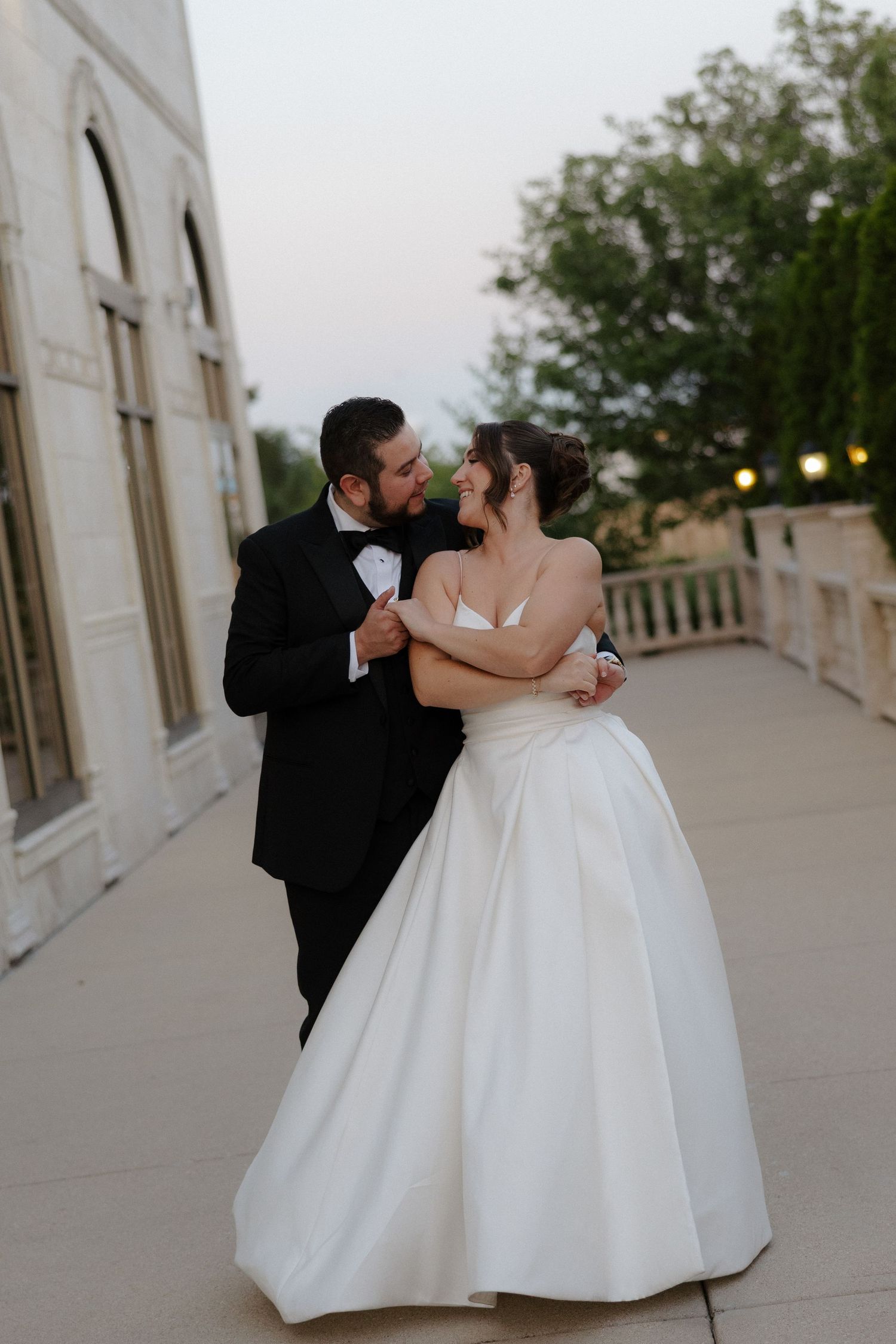 A bride and groom share a romantic dance outdoors at dusk, with the bride in a white ball gown and groom in a black tuxedo.
