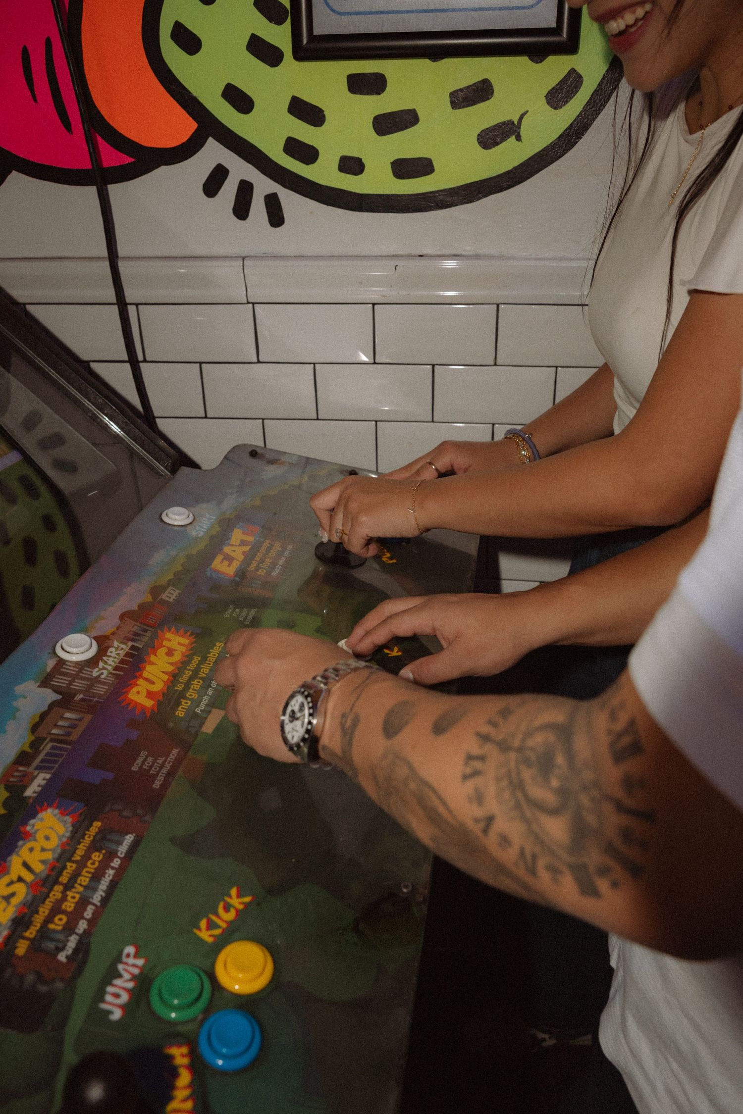 Two people playing a vintage arcade game with joystick controls, one arm featuring tattoos visible.