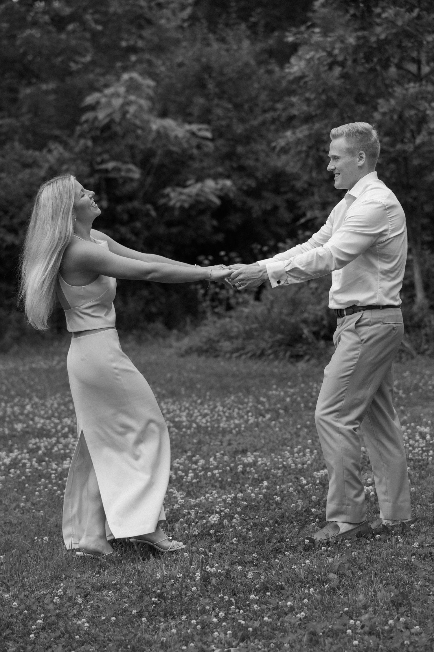 Black and white photo of a couple holding hands and dancing playfully in a grassy field with trees in the background.