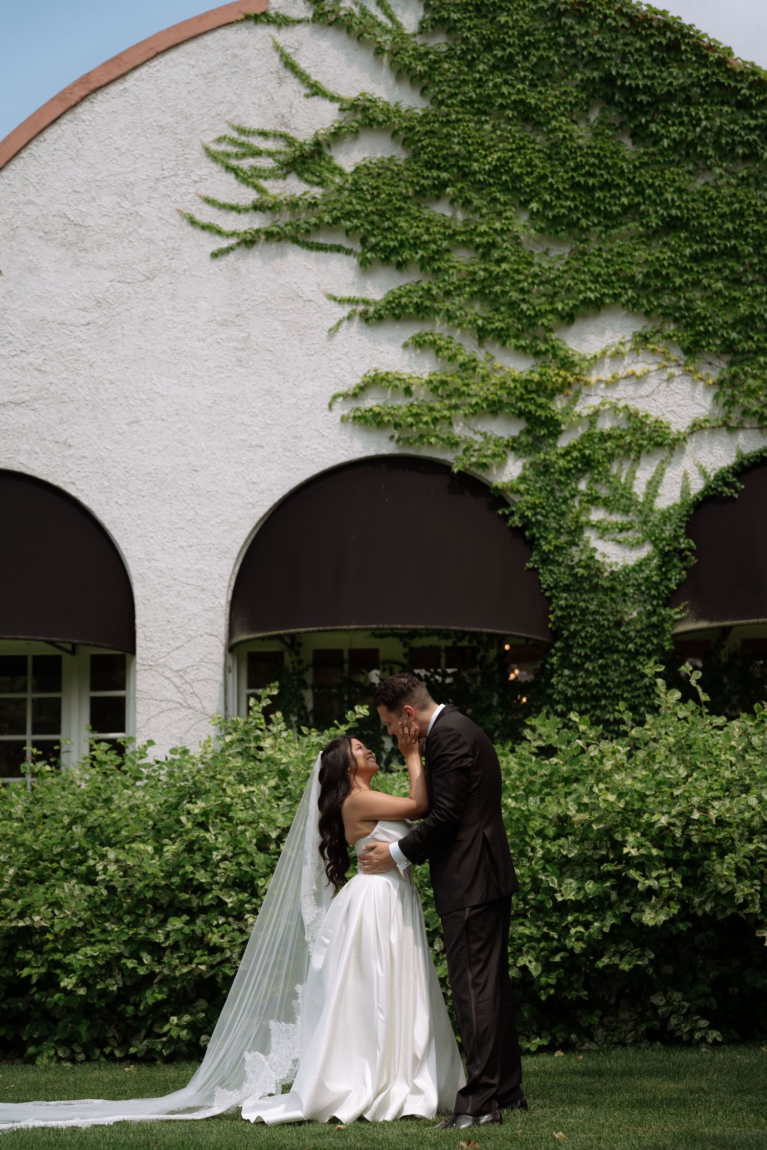 Bride and groom sharing a kiss in front of ivy-covered white building with arched windows.