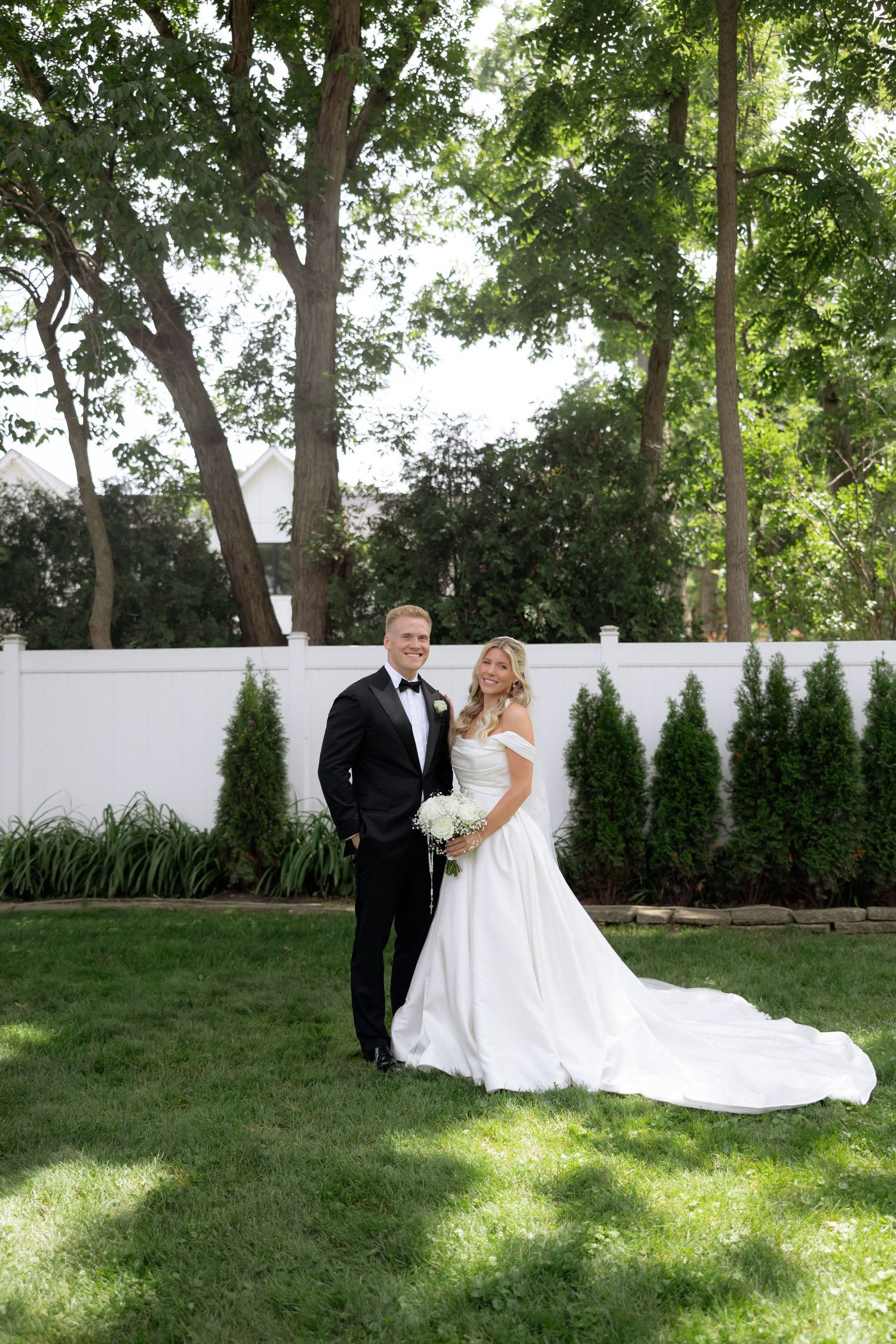 Bride and groom pose together on green lawn with white fence and tall trees in background.